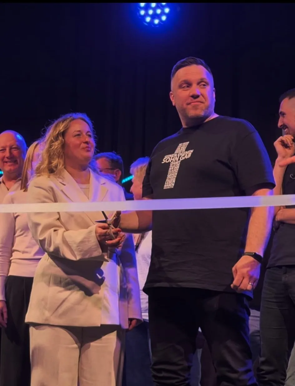 Ein Mann schneidet ein weißes Band mit einer Schere durch, umgeben von weiteren Personen. Im Hintergrund eine dunkle Bühne und Lichteffekte.