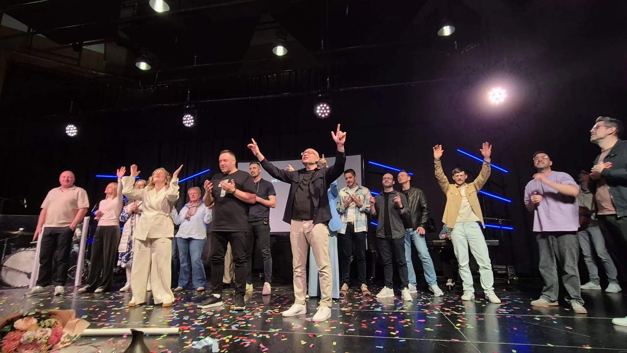 Eine Gruppe von etwa 20 Personen steht auf einer Bühne und hebt die Arme in die Luft. Im Hintergrund eine große Leinwand und Bühnenbeleuchtung.