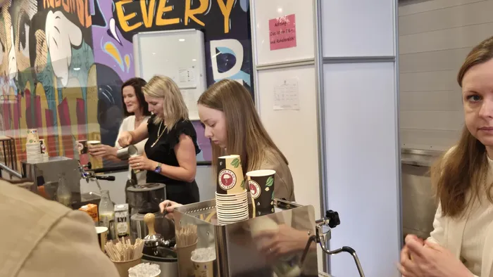 Zwei Frauen bedienen an einer mobilen Kaffee- und Eistheke, während eine weitere Person im Hintergrund steht. Im Hintergrund ein farbenfrohes Wandbild und ein Hinweis auf Eis mit EU-Zutaten.
