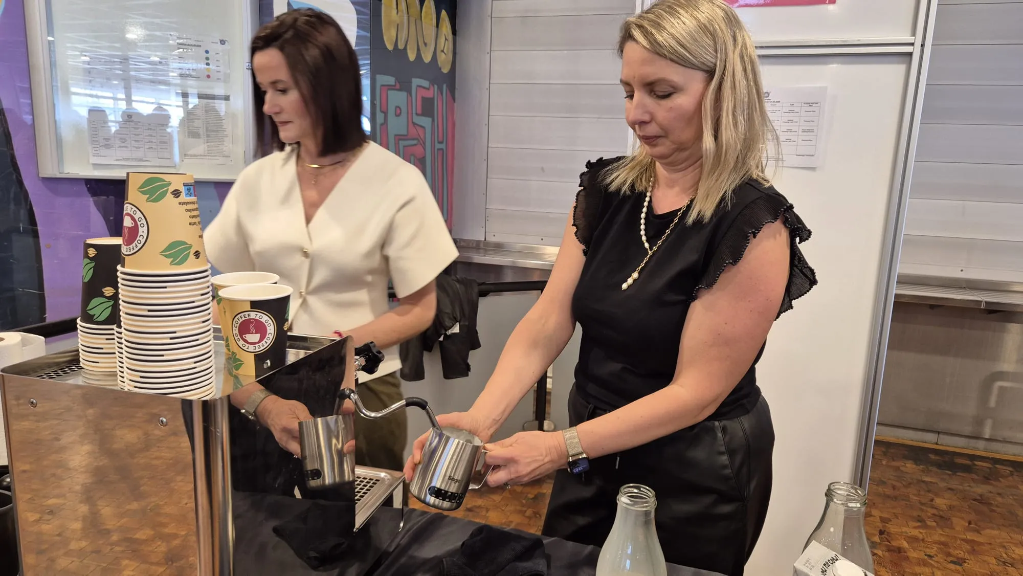 Zwei Frauen stehen hinter einer Espressomaschine und bereiten Kaffee zu. Im Hintergrund sind weitere Personen und ein Raum mit Tischen und Stühlen zu sehen.