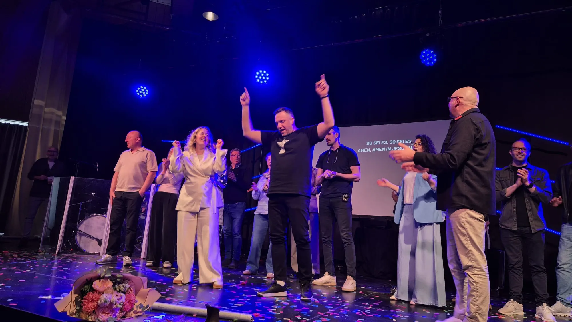 Eine Gruppe von Menschen steht auf einer Bühne vor einer großen Leinwand mit Text, einige heben die Arme. Im Hintergrund ist ein Publikum in einem großen Saal zu sehen.
