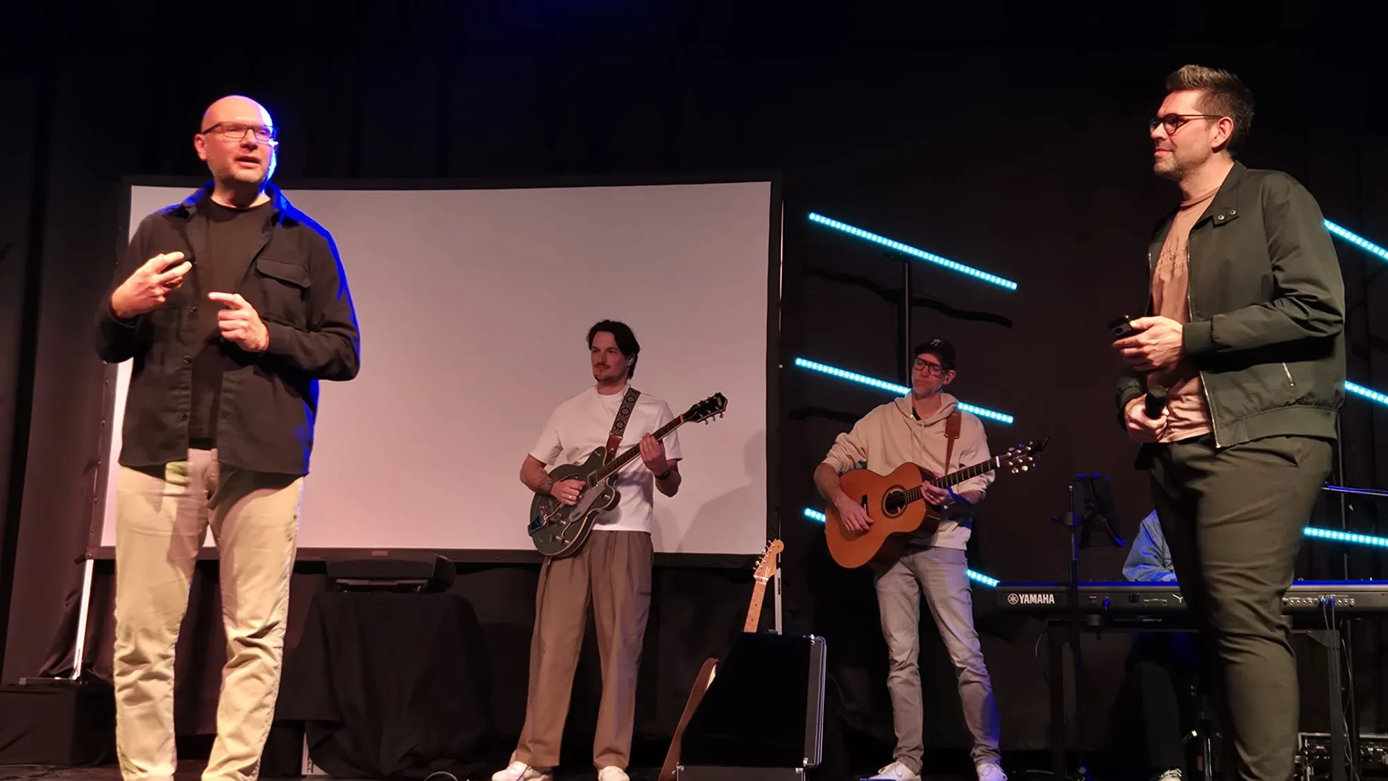 Vier Männer auf einer Bühne, zwei von ihnen spielen Gitarre, während die anderen sprechen. Im Hintergrund eine große weiße Leinwand und blaue Lichtelemente.