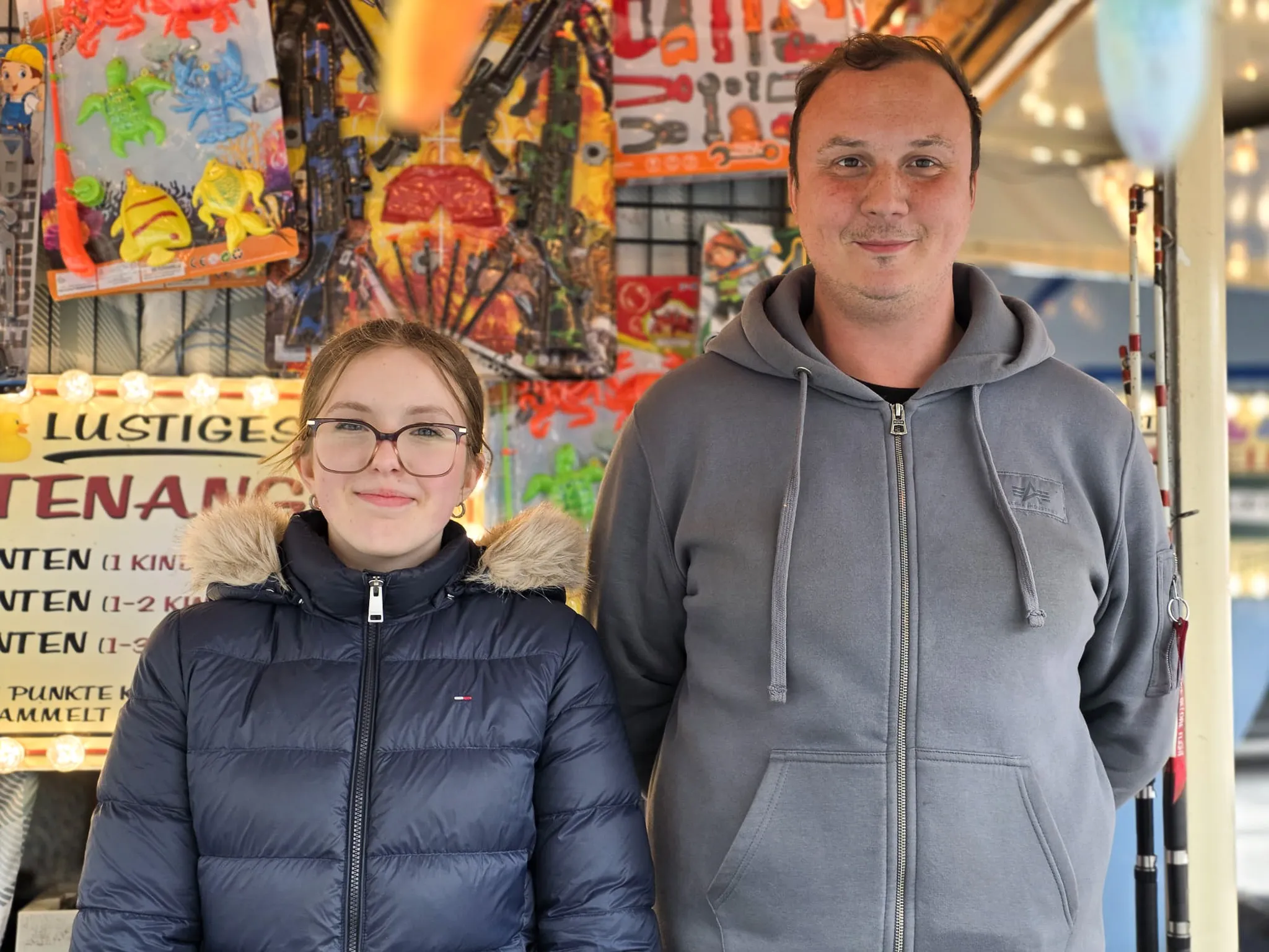 Ein Mann und ein Mädchen stehen vor einem Kirmesstand mit Spielzeug und Süßigkeiten. Im Hintergrund sind weitere Kirmesbuden und Lichter zu sehen.