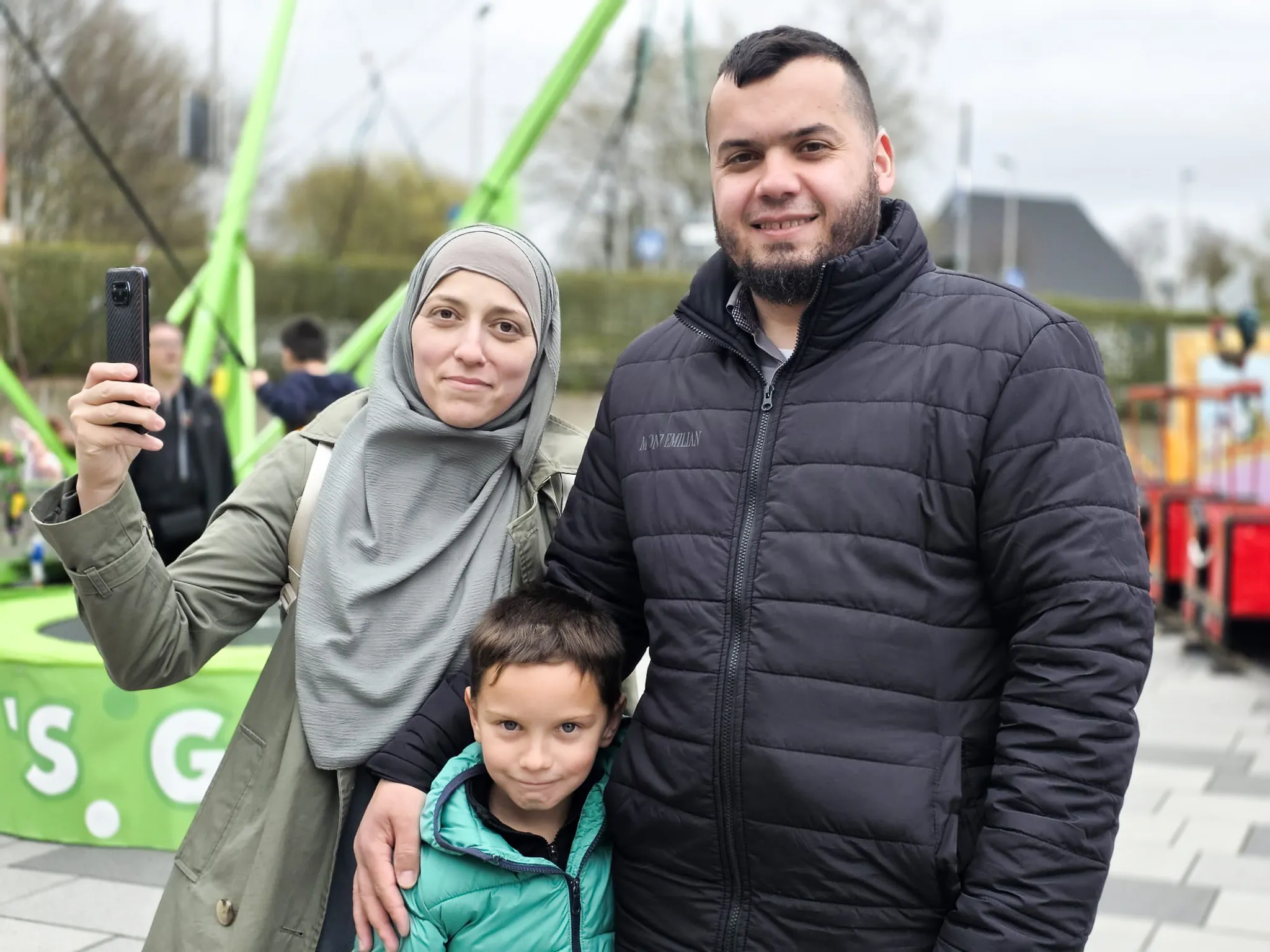 Familie mit Kind macht ein Selfie vor dem Hintergrund einer Kirmesattraktion.