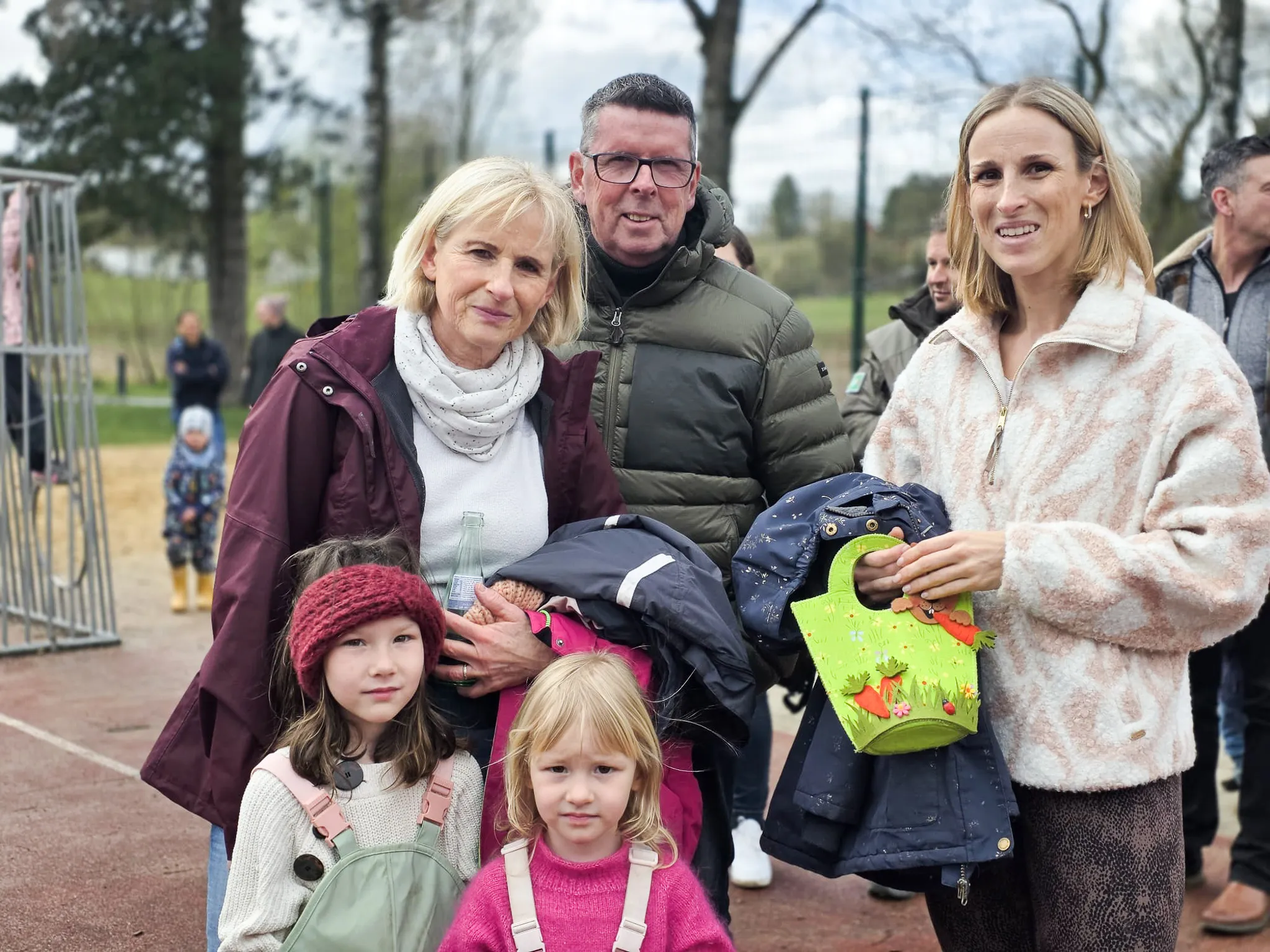 Familie mit zwei kleinen Mädchen vor dem Hintergrund einer Ostereiersuche im Freien. Eine Frau hält einen kleinen Osterkorb.