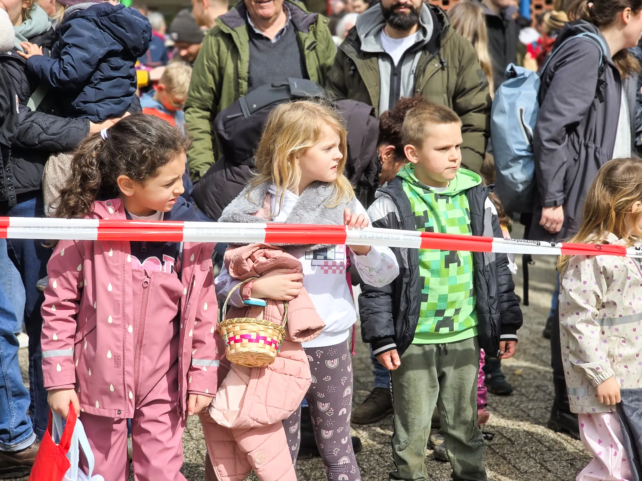 Drei Kinder stehen vor einer Absperrung, die Körbe mit Ostereiern halten. Im Hintergrund sind weitere Menschen zu sehen.