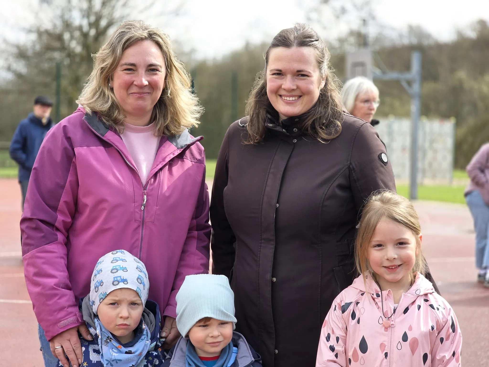 Zwei Frauen und drei Kinder stehen lächelnd vor einer Sportfläche. Die Kinder tragen Winterjacken und Mützen.