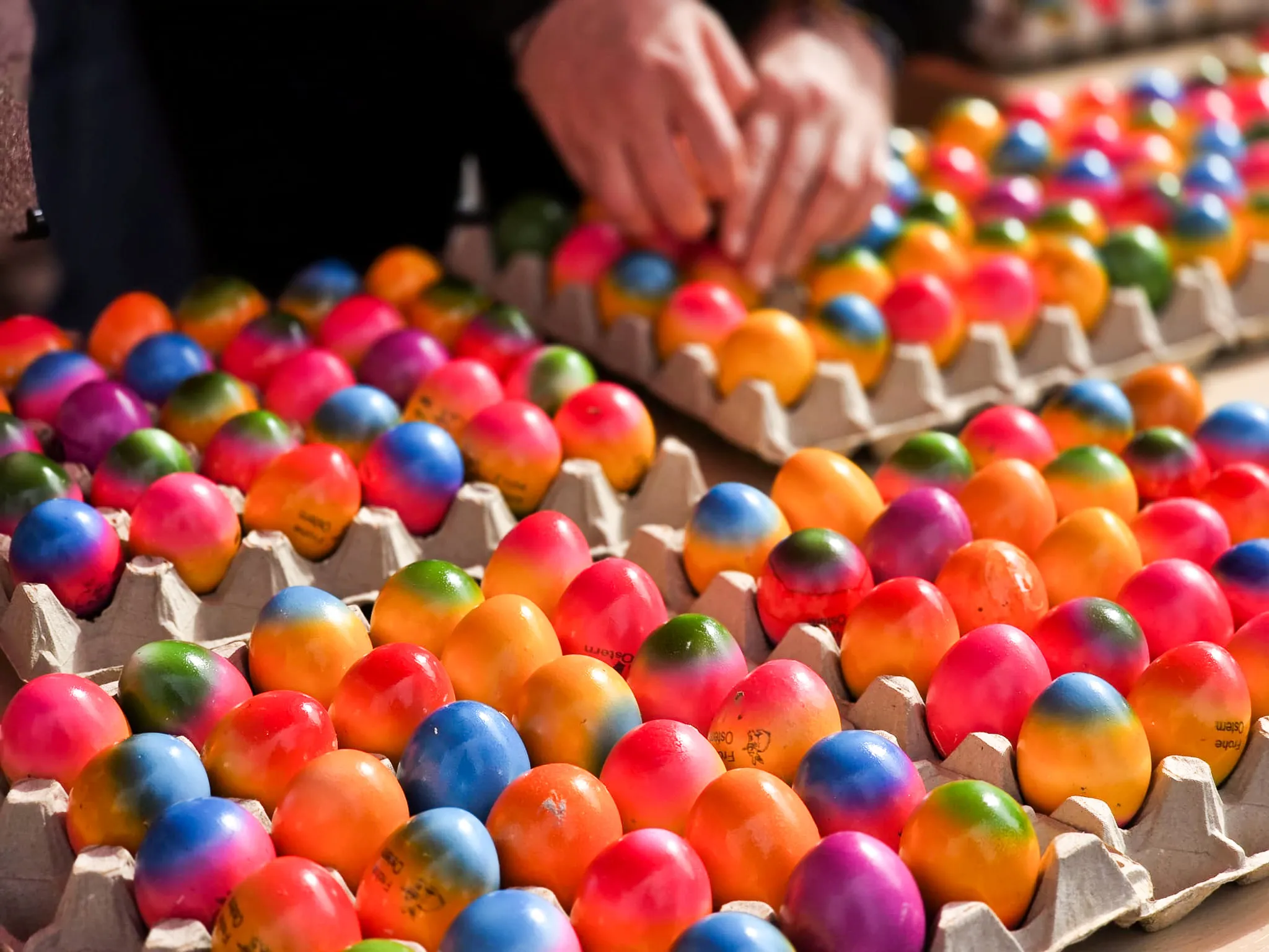 Bunte Ostereier in einem Eierkarton, über denen eine Hand nach weiteren Eiern greift.