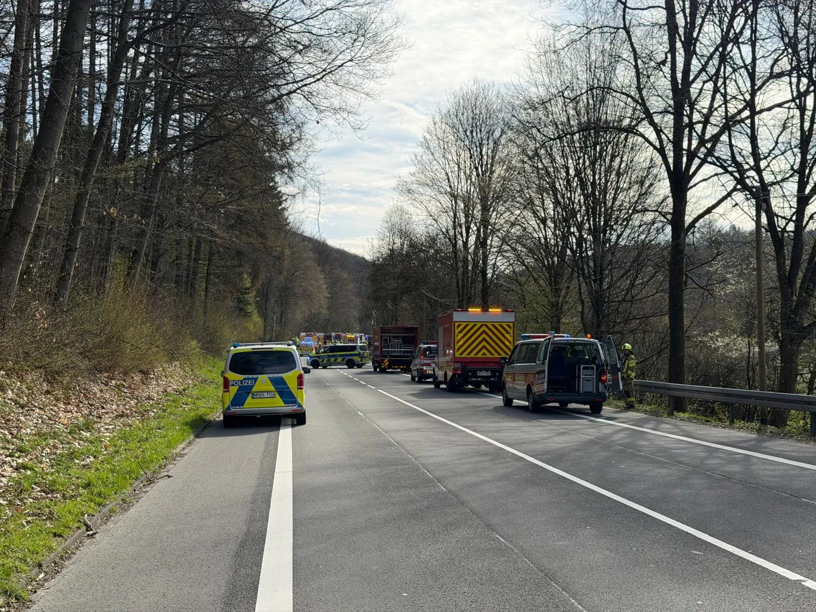 Polizeifahrzeuge und Einsatzkräfte sichern eine Vollsperrung auf einer Landstraße zwischen Bäumen, nachdem es zu einem Verkehrsunfall kam.