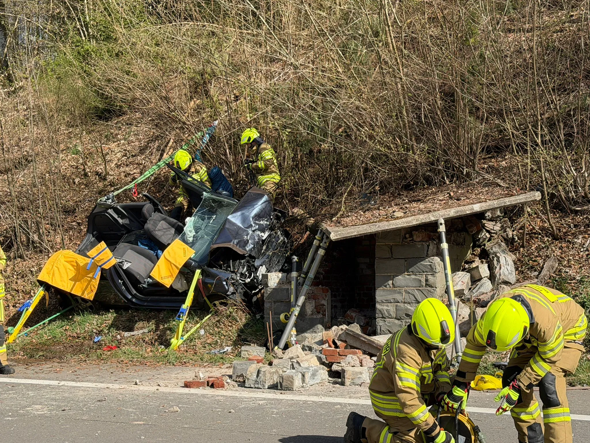Ein stark beschädigtes Auto ist in ein Gebäude an einer Landstraße eingekracht. Feuerwehrleute sichern die Unfallstelle und arbeiten neben dem Wrack.