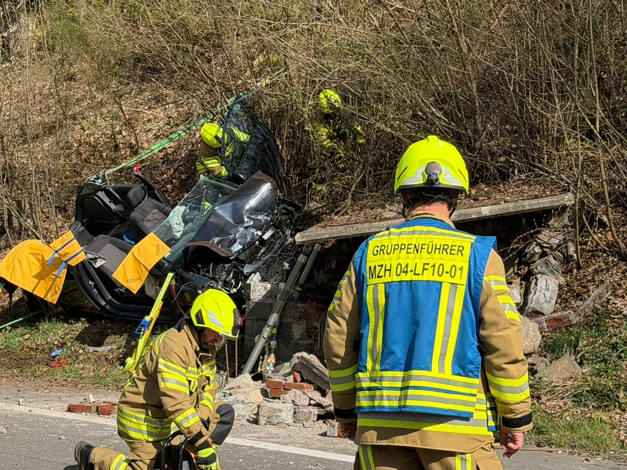 Feuerwehrleute stehen an einem Unfallort, wo ein Auto schwer beschädigt in einem Abhang liegt. Im Hintergrund sind weitere Einsatzkräfte zu sehen.