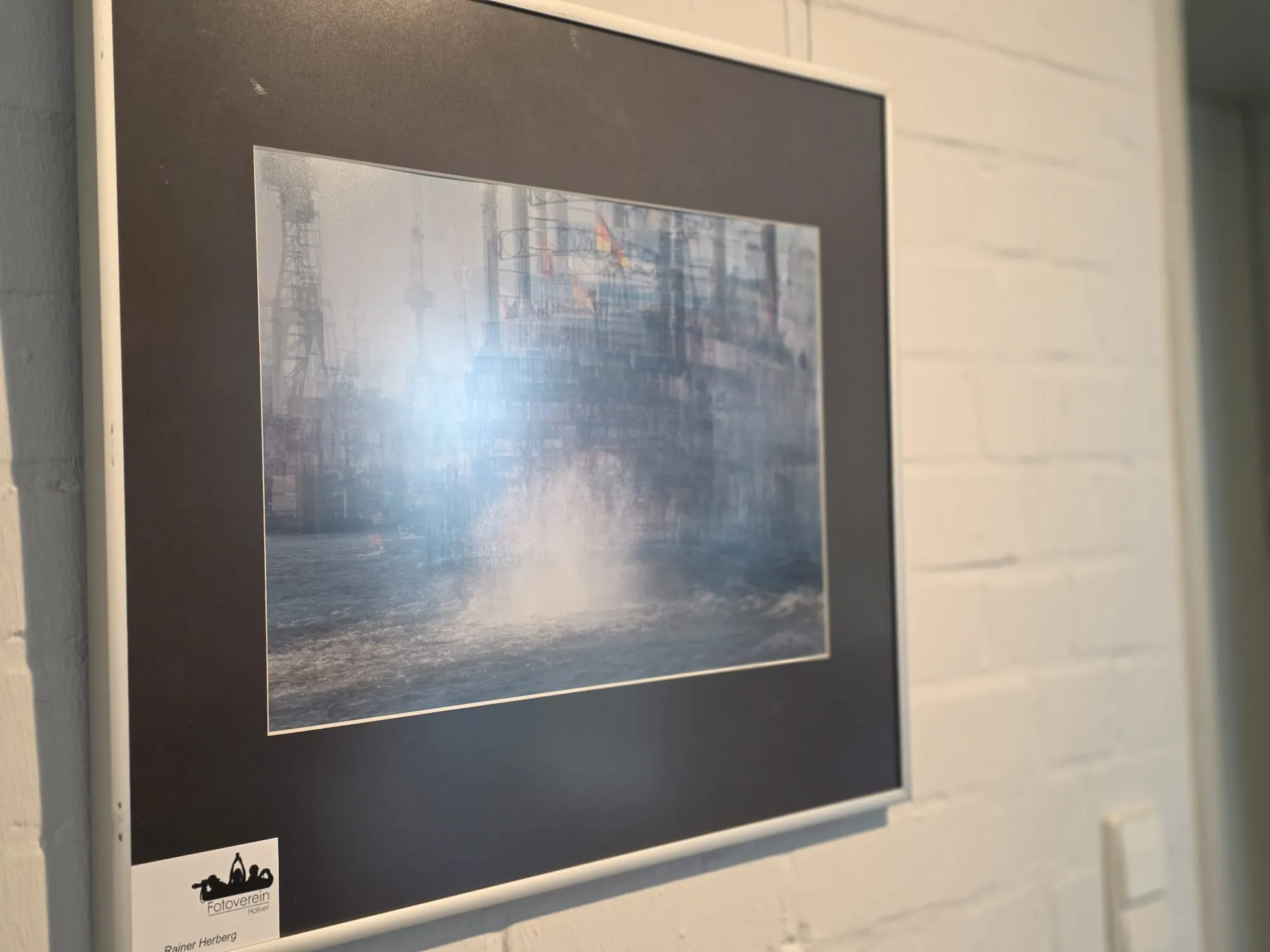 Schwarz-Weiß-Foto einer Wassersprenge vor einer Industrielandschaft mit Türmen und Rohren.