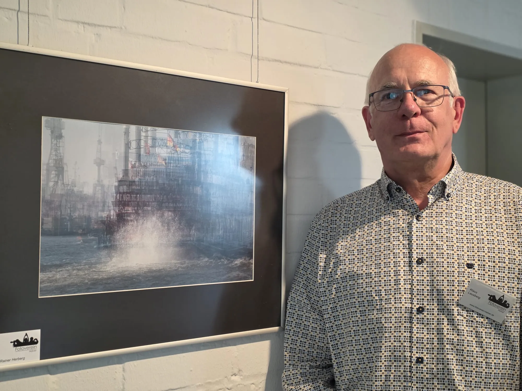 Ein Mann steht neben einem gerahmten Schwarzweißfoto einer stürmischen See mit einer Mole im Vordergrund. Das Foto zeigt hohe Wellen und eine Gischt, die an der Mole hochsprüht.