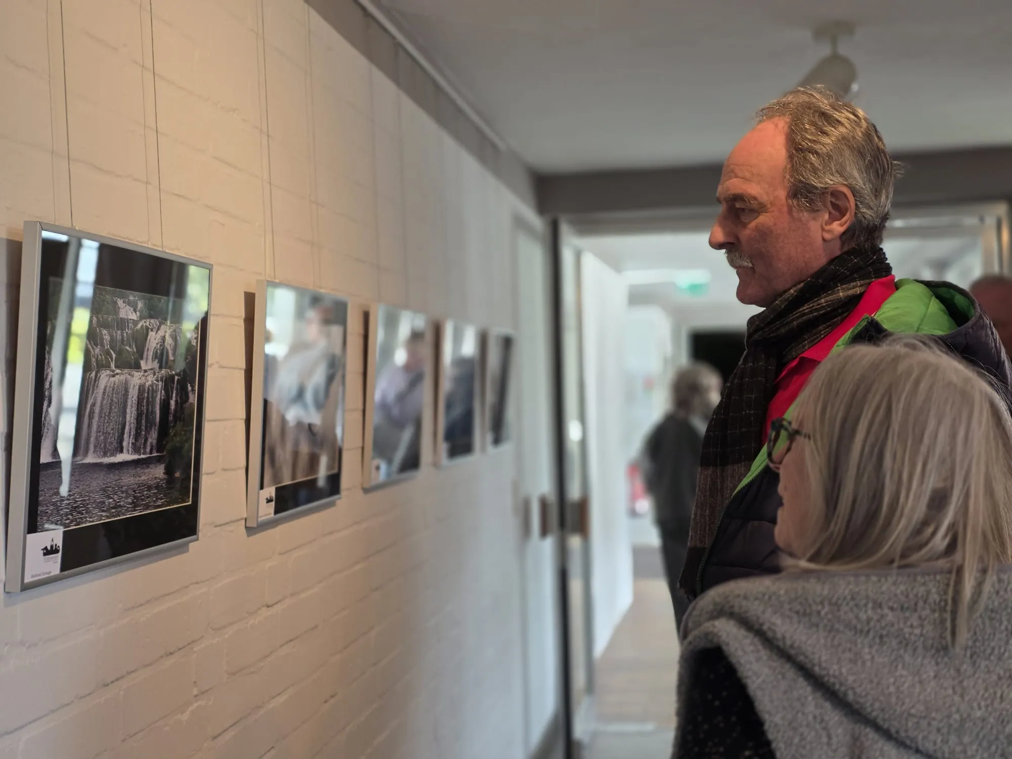 Zwei Besucher betrachten eine Reihe gerahmter Schwarzweißfotografien an einer weißen Backsteinwand, die Wasserfälle und andere Wasserszenen zeigen.
