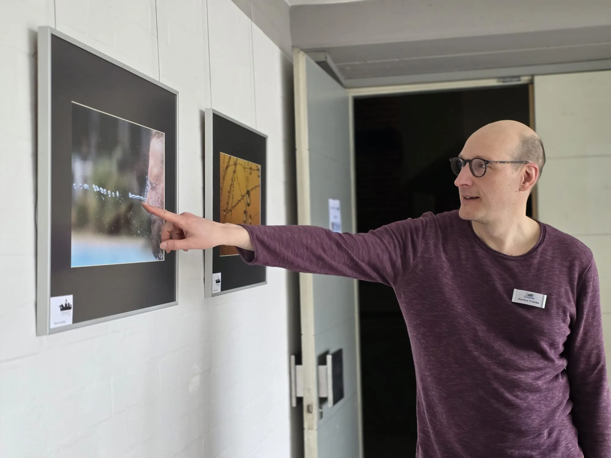 Ein Mann mit Brille zeigt auf ein gerahmtes Foto an einer hellen Wand, neben dem ein weiteres Bild hängt. Die Ausstellung zeigt Fotografien zum Thema Wasser.