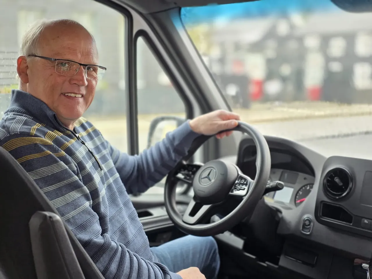 Ein älterer Mann mit Brille lächelt und hält das Lenkrad eines grauen Busses fest.