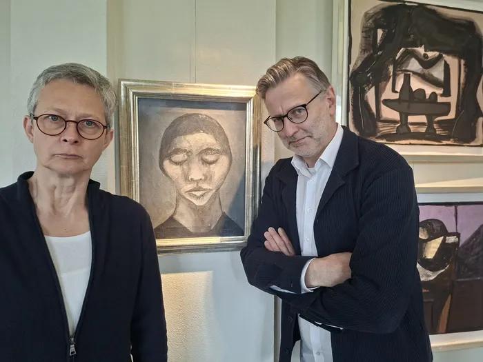 Zwei Personen, eine Frau mit kurzem grauem Haar und ein Mann mit dunklem Haar, stehen vor Gemälden in einem Innenraum. Im Hintergrund ist ein Porträt eines Mannes mit Mütze zu sehen.