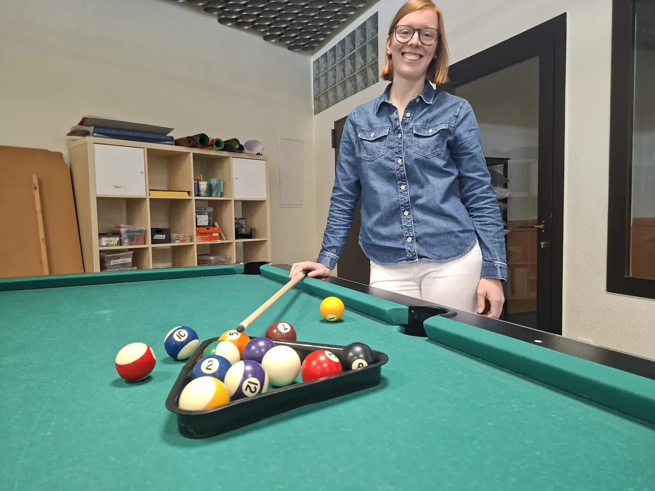 Jannica Beckmann steht lächelnd neben einem Billardtisch und hält einen Queue bereit. Im Hintergrund sind Regale mit Spielmaterialien zu sehen.