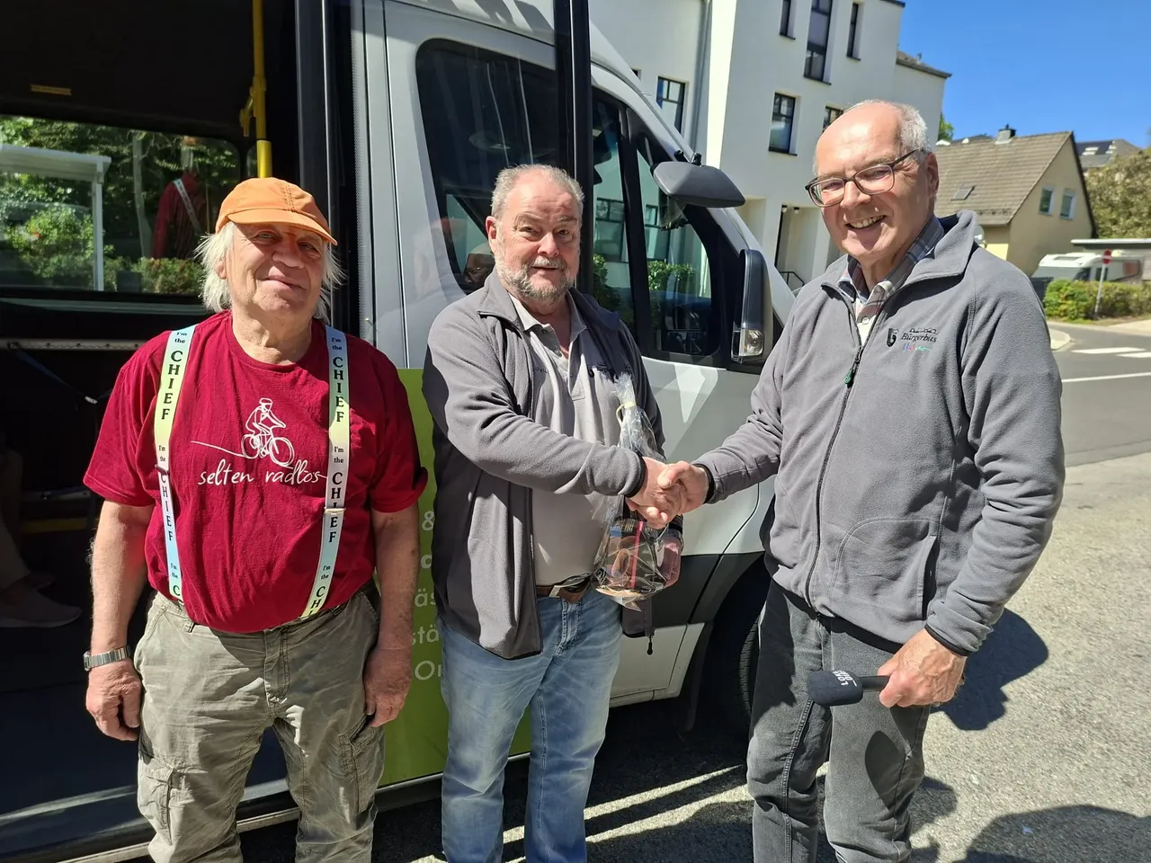 Drei Männer schütteln sich vor einem weißen Kleinbus die Hände. Der Mann links trägt ein rotes T-Shirt mit Aufschrift, der Mann in der Mitte ein graues Hemd und der Mann rechts eine graue Jacke.