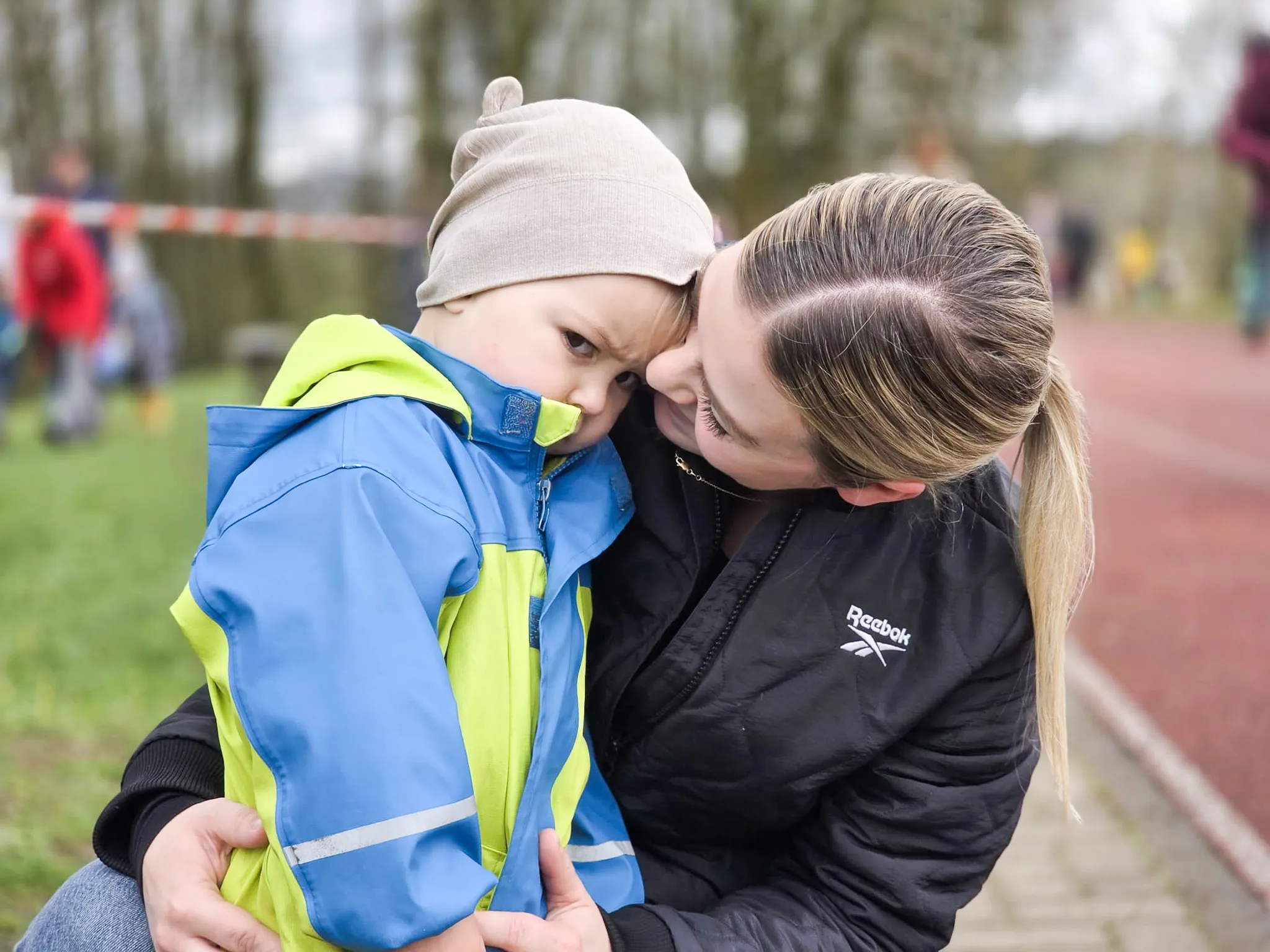 Eine Frau küsst einen kleinen Jungen auf die Stirn, während sie ihn hält. Beide stehen auf einer grünen Wiese, im Hintergrund sind weitere Personen und ein roter Laufbahn zu sehen.