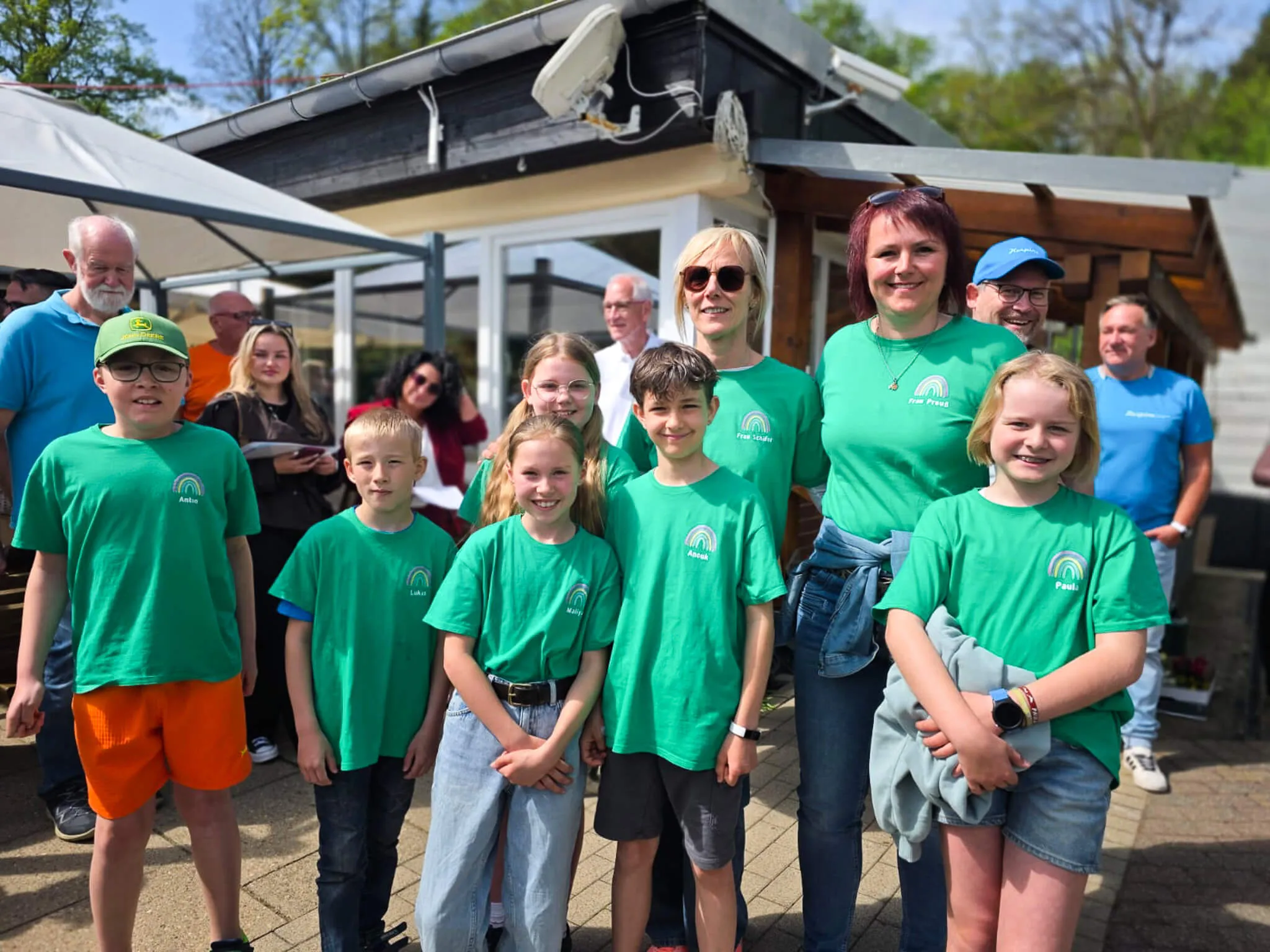 Eine Gruppe von Kindern und Erwachsenen in grünen T-Shirts mit Regenbogenmotiv lächelt in die Kamera. Im Hintergrund ein Gebäude und weitere Personen.