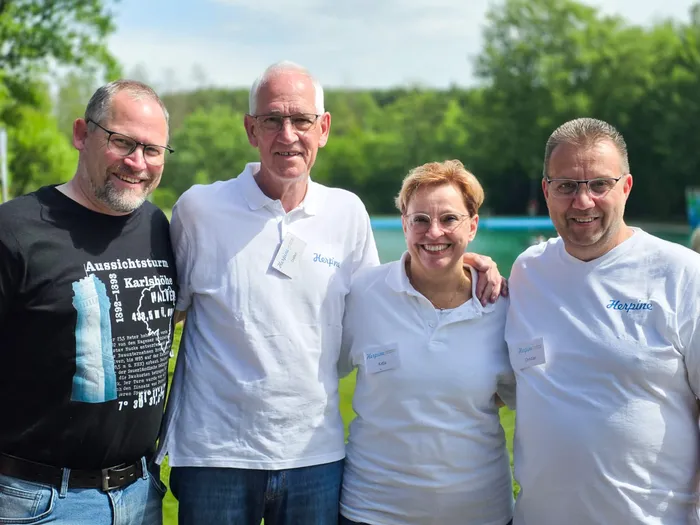 Fünf lächelnde Menschen stehen im Freien vor einer grünen Hecke. Sie tragen Namensschilder mit der Aufschrift „Herpine“ und weiße oder helle Kleidung.
