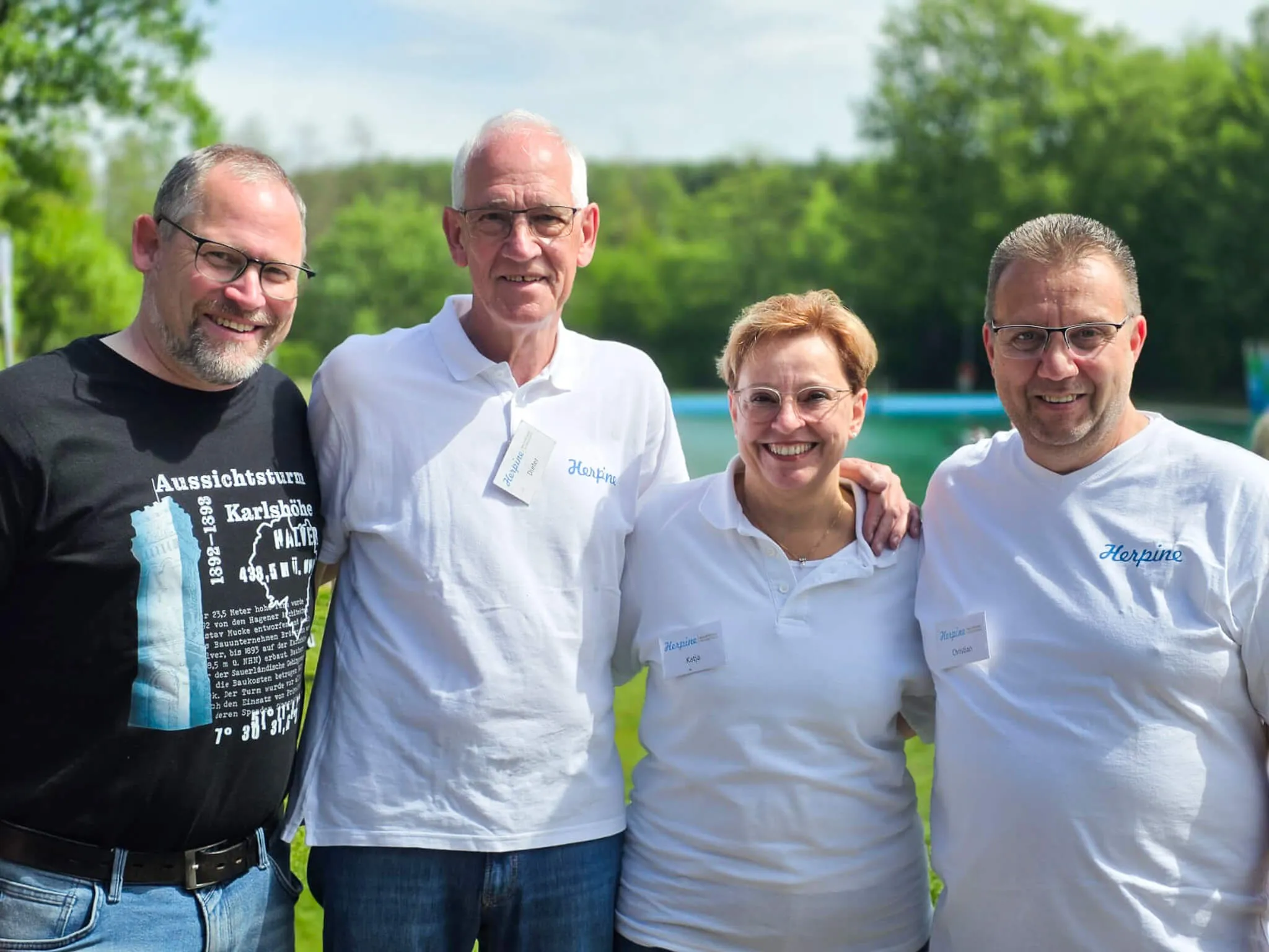 Fünf lächelnde Menschen stehen im Freien vor einer grünen Hecke. Sie tragen Namensschilder mit der Aufschrift „Herpine“ und weiße oder helle Kleidung.