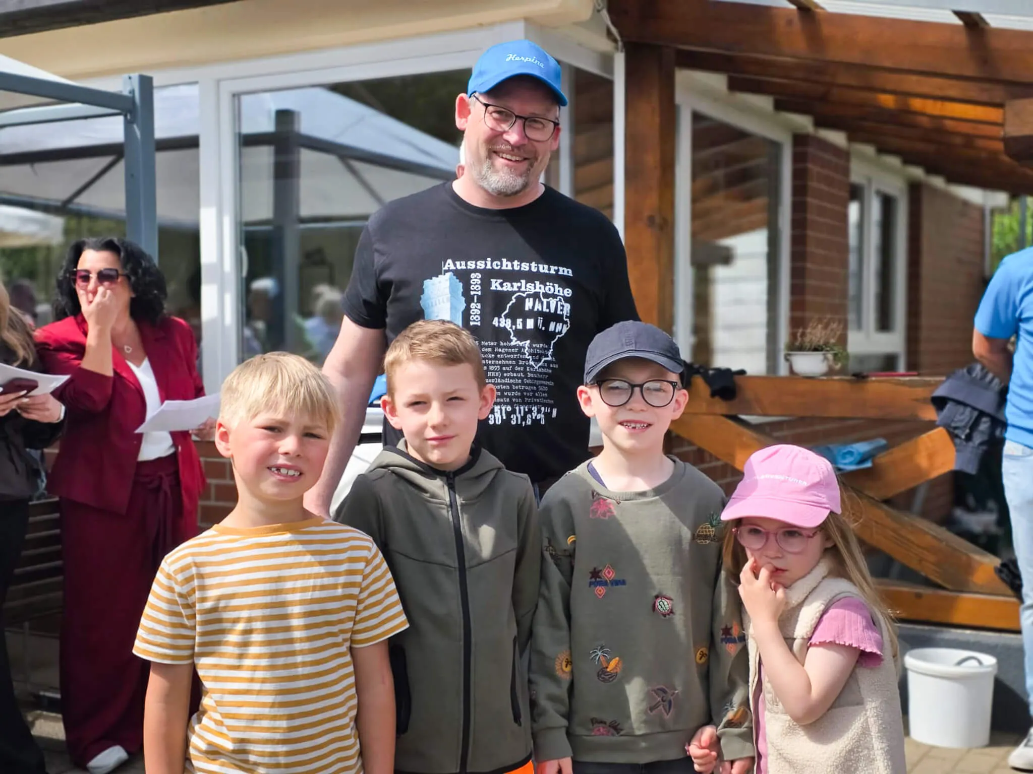 Vier Jungen stehen vor einem Mann mit einem bedruckten T-Shirt im Freien. Im Hintergrund ist ein Gebäude mit einer Markise und Bäume zu sehen.