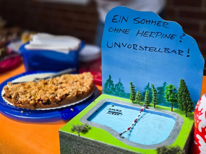 Ein selbstgemachtes Schild mit der Aufschrift „Ein Sommer ohne Herpine? Unvorstellbar!“ steht vor einer Miniaturlandschaft mit Schwimmbad und Bäumen, daneben ein Stück Kuchen und ein Becher mit Eis.