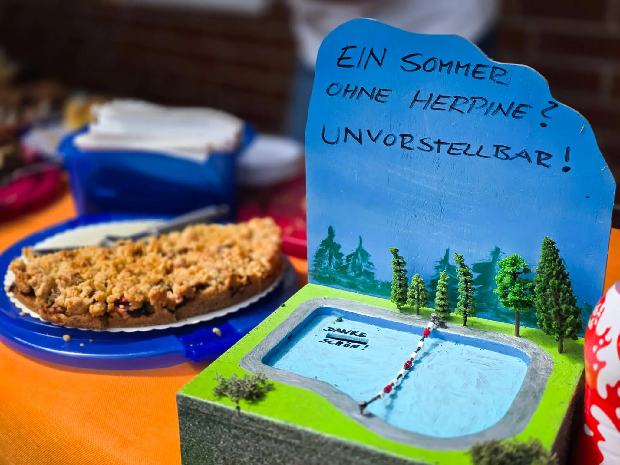 Ein selbstgemachtes Schild mit der Aufschrift „Ein Sommer ohne Herpine? Unvorstellbar!“ steht vor einer Miniaturlandschaft mit Schwimmbad und Bäumen, daneben ein Stück Kuchen und ein Becher mit Eis.