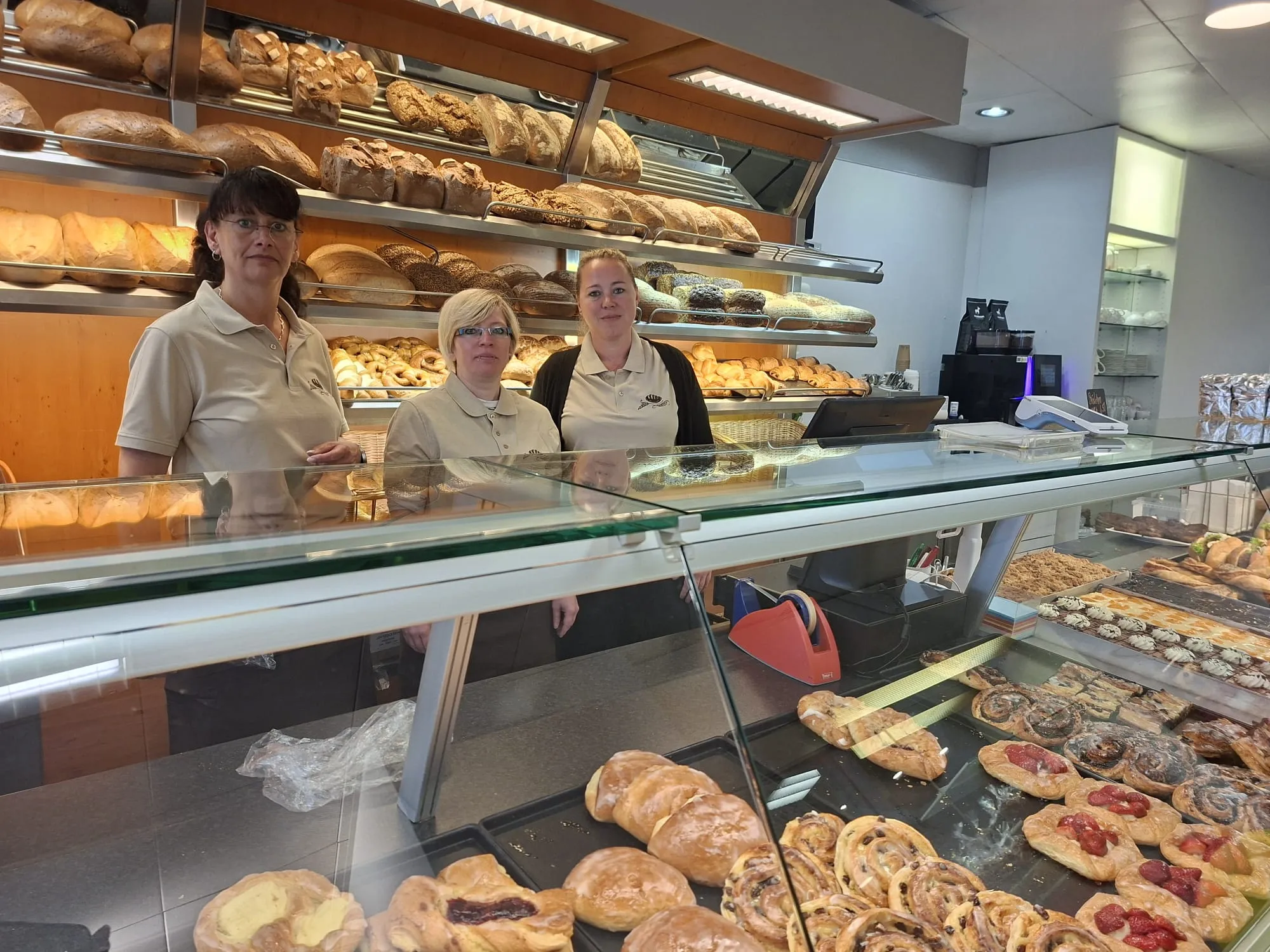 Drei Verkäuferinnen stehen hinter einer Theke mit einer großen Auswahl an Gebäck und Brot. Im Hintergrund sind Regale mit weiteren Backwaren zu sehen.