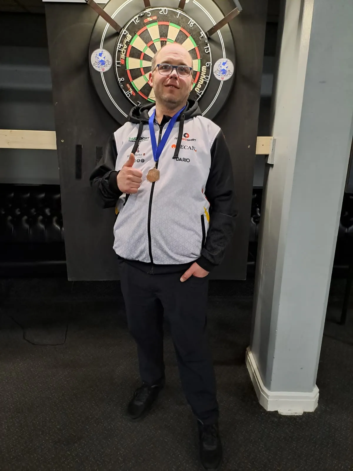 Dario Gentilcore, ein Mann mit Brille, trägt eine Jacke mit Aufschrift „Dario“ und eine Bronzemedaille um den Hals. Im Hintergrund ist eine Dartscheibe zu sehen.