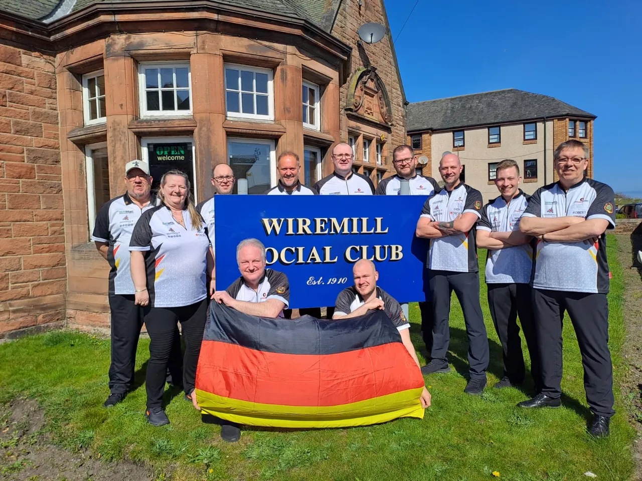 Gruppe von Menschen in Vereinskleidung vor einem Gebäude mit dem Banner „Wiremill Social Club Est. 1910“. Ein Mann hält die Deutschlandflagge.