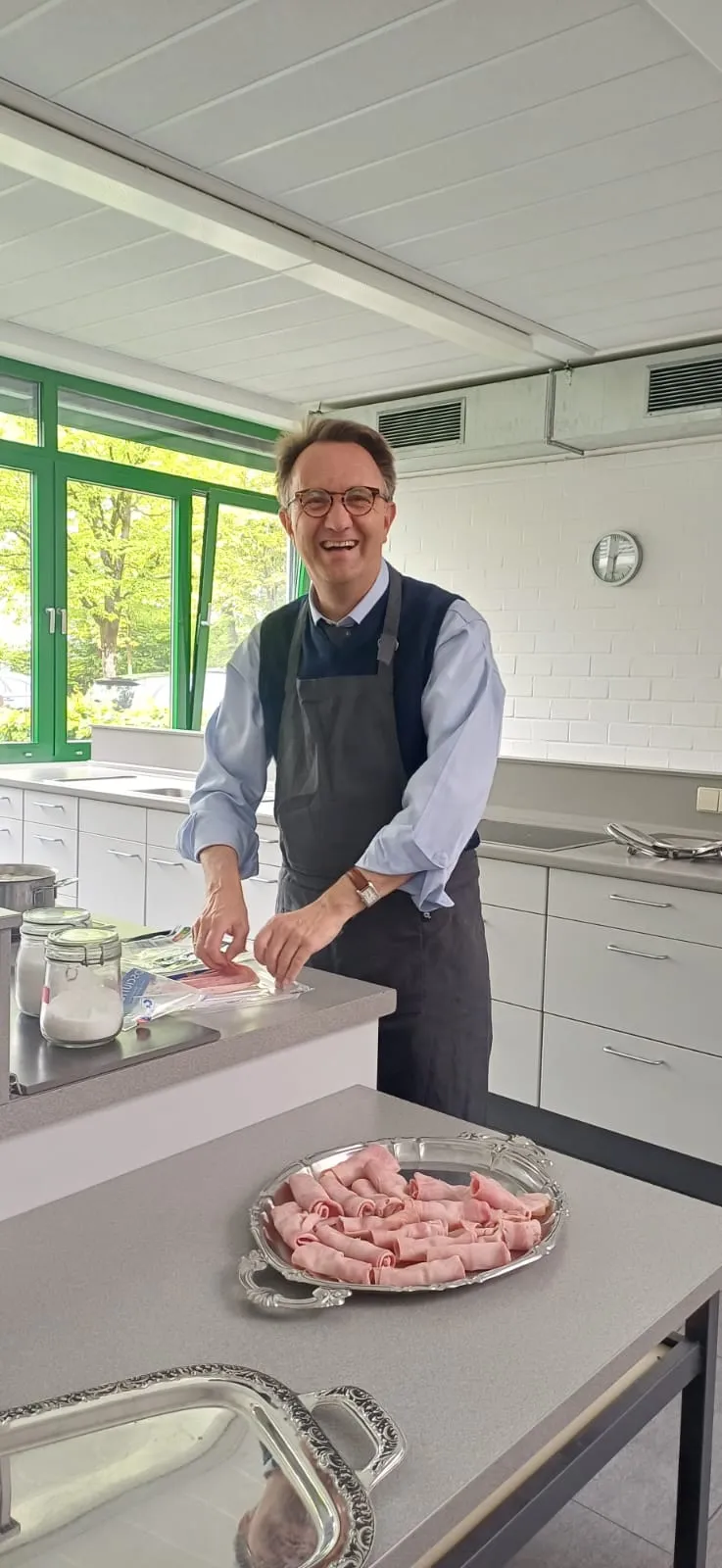 Schulleiter Paul Meurer steht in einer Küche und schneidet Aufschnitt auf einer Arbeitsfläche. Vor ihm liegt eine Platte mit bereits geschnittenen Scheiben.