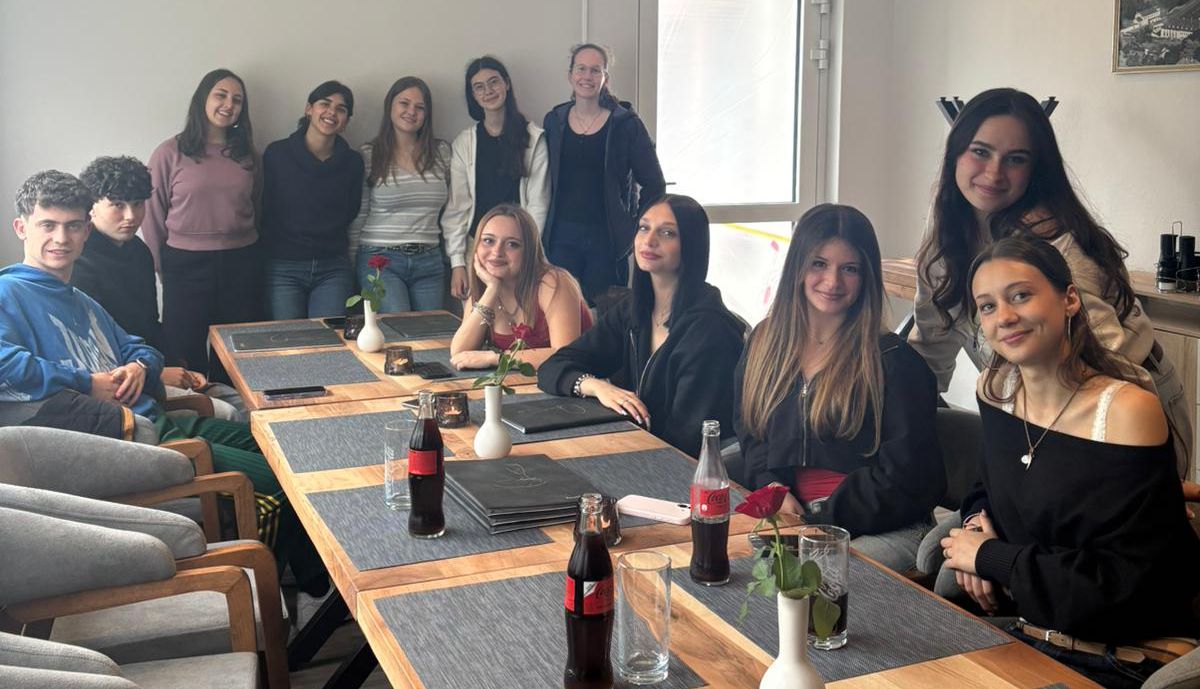 Gruppe von Jugendlichen sitzt um einen Tisch mit Getränken und einer einzelnen roten Rose in einer hellen Umgebung. Das Bild zeigt einen Schüleraustausch zwischen dem AFG und dem Instituto Soleri Bertoni aus Italien.