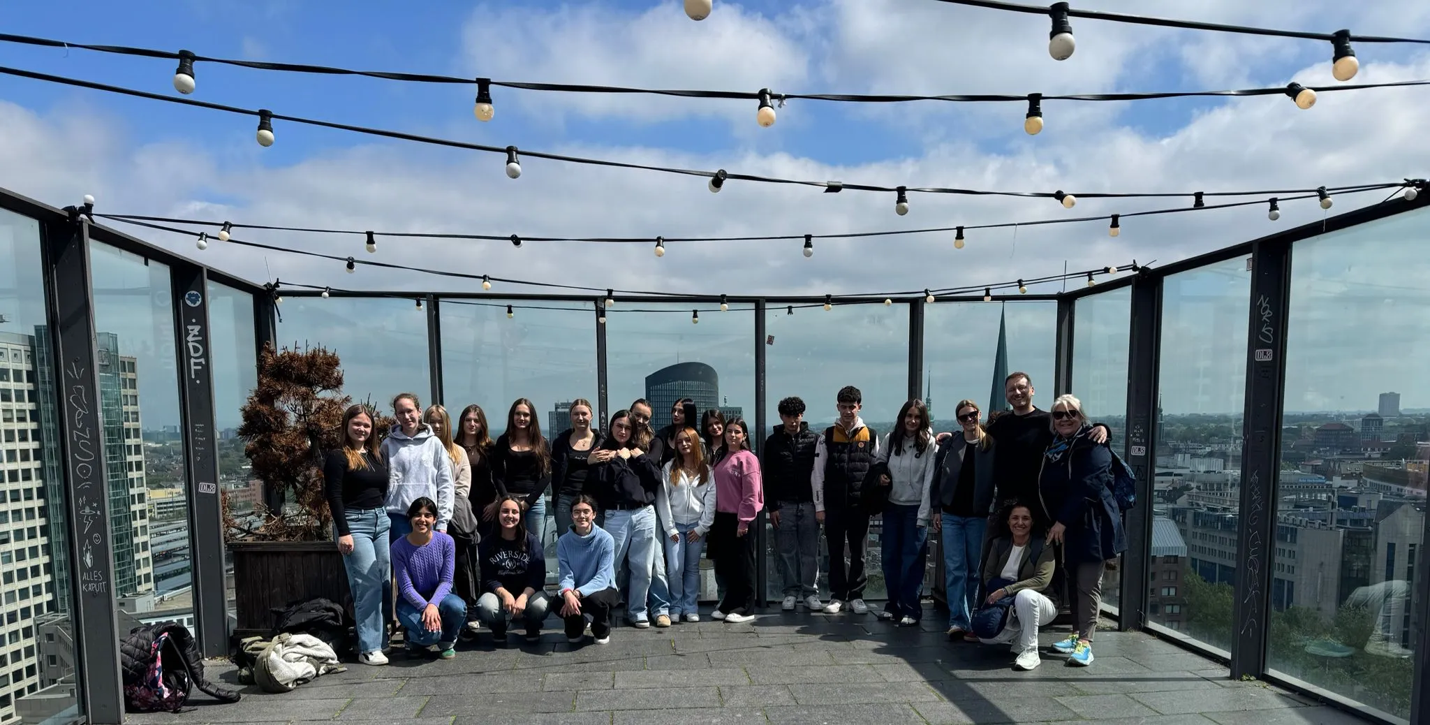 Eine Gruppe junger Menschen steht lächelnd auf einer Dachterrasse mit Glaswänden und Blick auf die Stadtlandschaft von Dortmund.