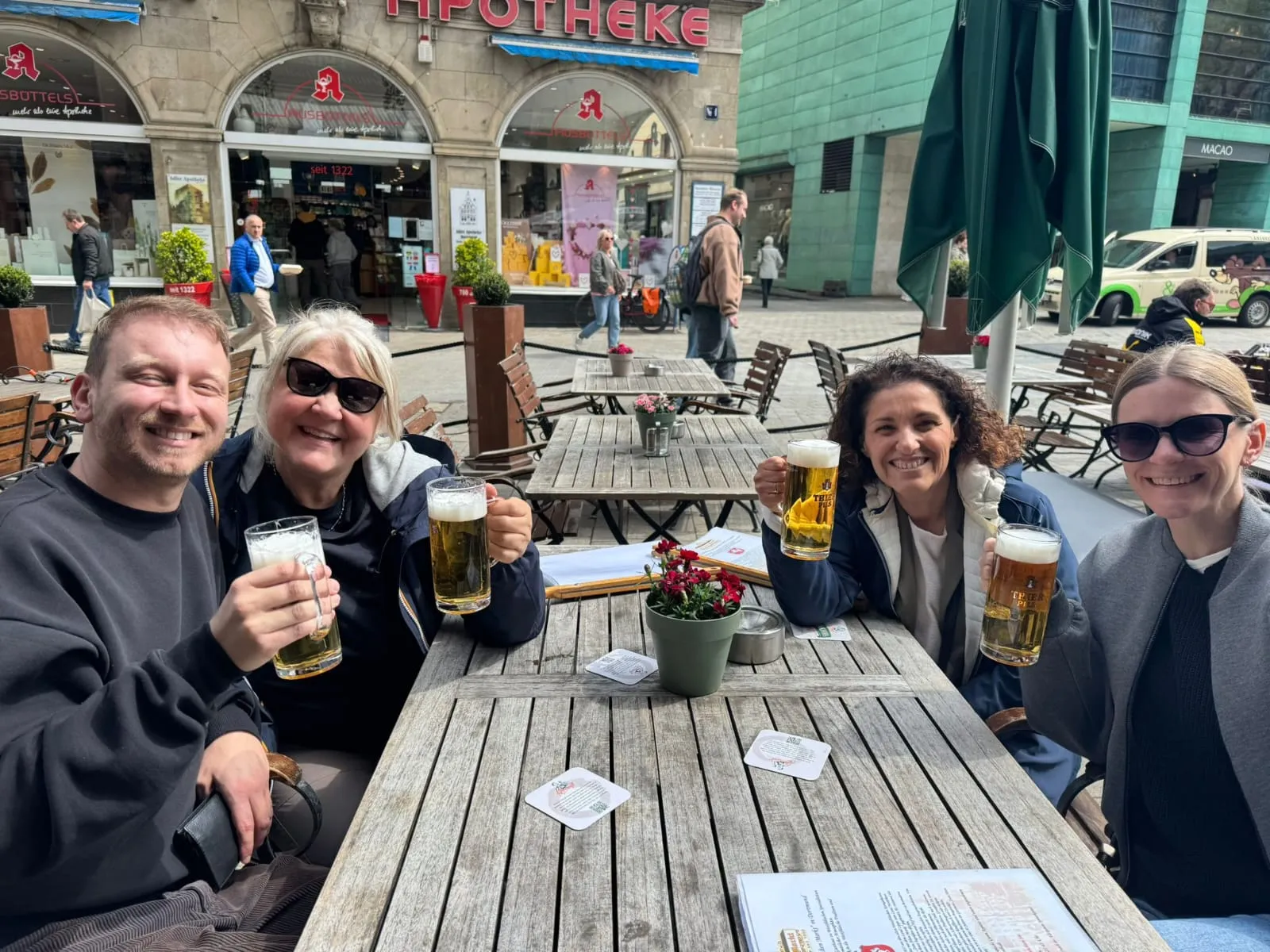 Vier Personen sitzen an einem Holztisch im Freien und heben jeweils ein Glas Bier. Im Hintergrund ist eine Straßenszene mit Geschäften und Passanten zu sehen.