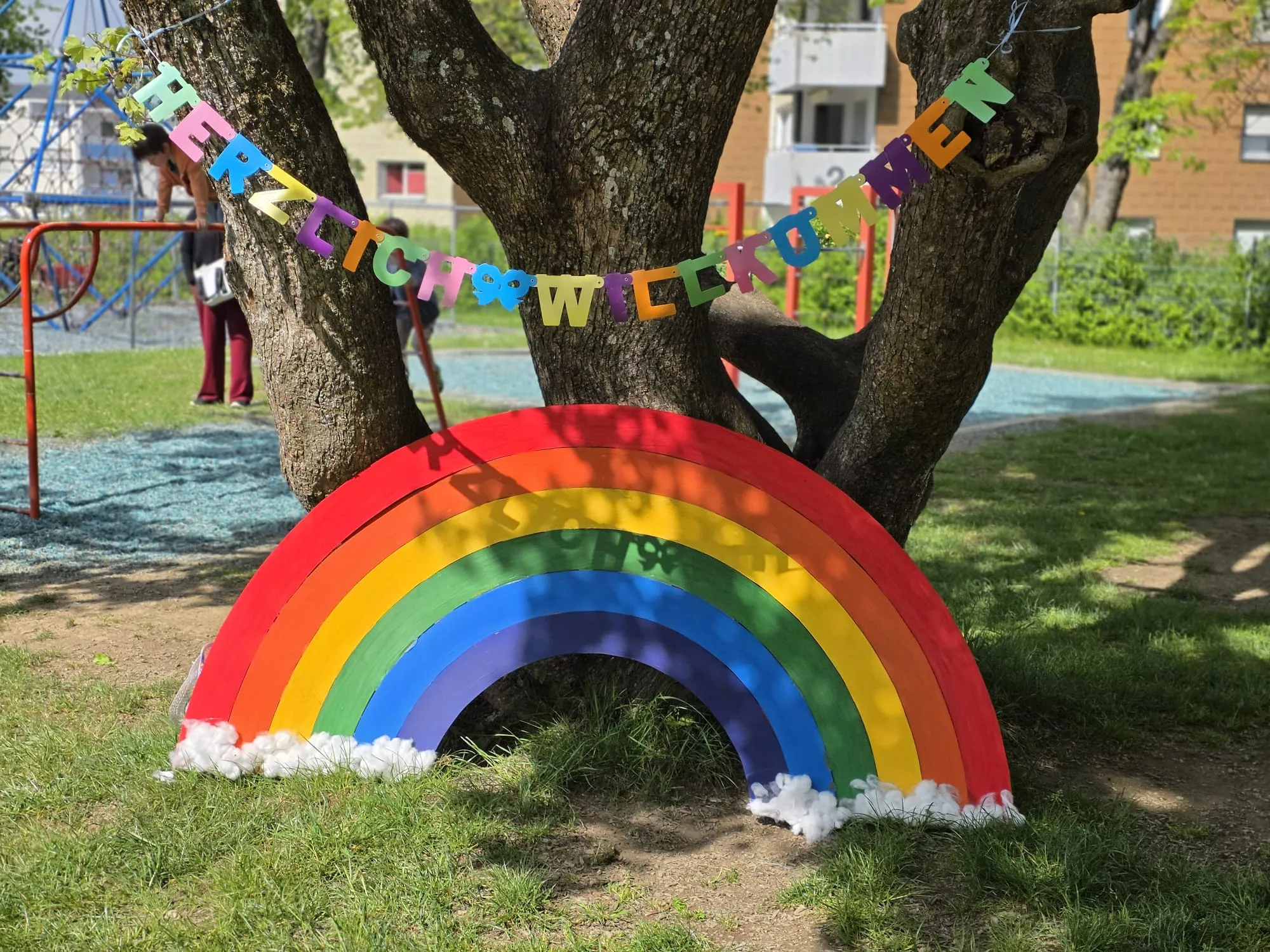 Ein großes, buntes Regenbogen-Holzmodell steht vor einem Baum auf einem Schulhof, umgeben von Kindern und einer Girlande mit der Aufschrift „Herzlich Willkommen“.