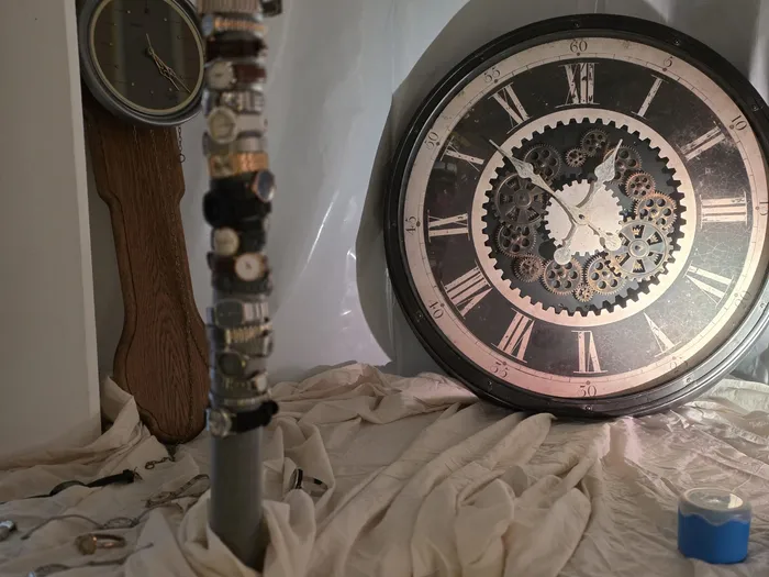 Zwei Uhren, eine Standuhr und eine Wanduhr mit freiliegendem Uhrwerk, liegen auf einer weißen Stoffbahn. Im Hintergrund ist eine weiße Wand zu sehen.