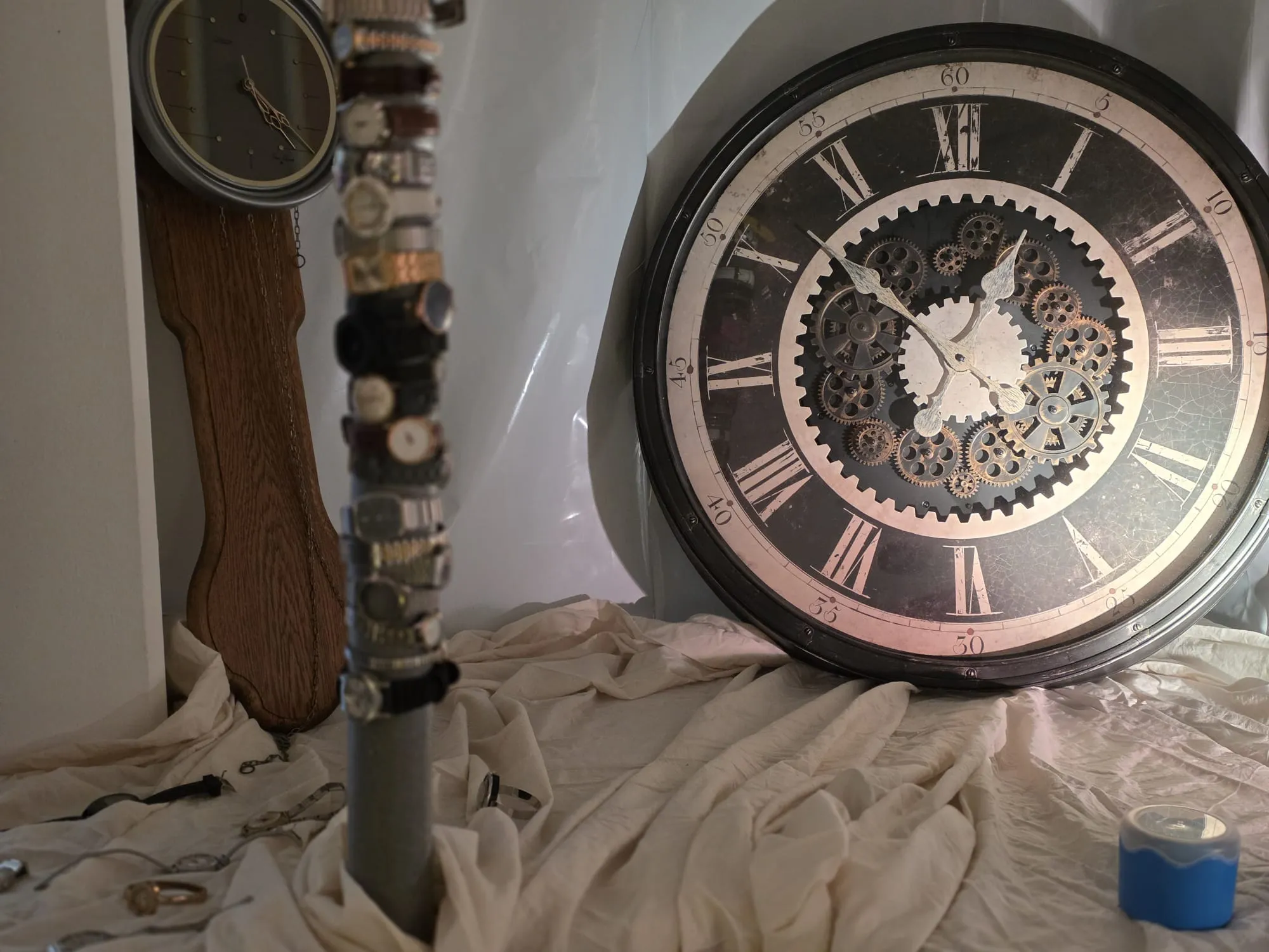 Zwei Uhren, eine Standuhr und eine Wanduhr mit freiliegendem Uhrwerk, liegen auf einer weißen Stoffbahn. Im Hintergrund ist eine weiße Wand zu sehen.