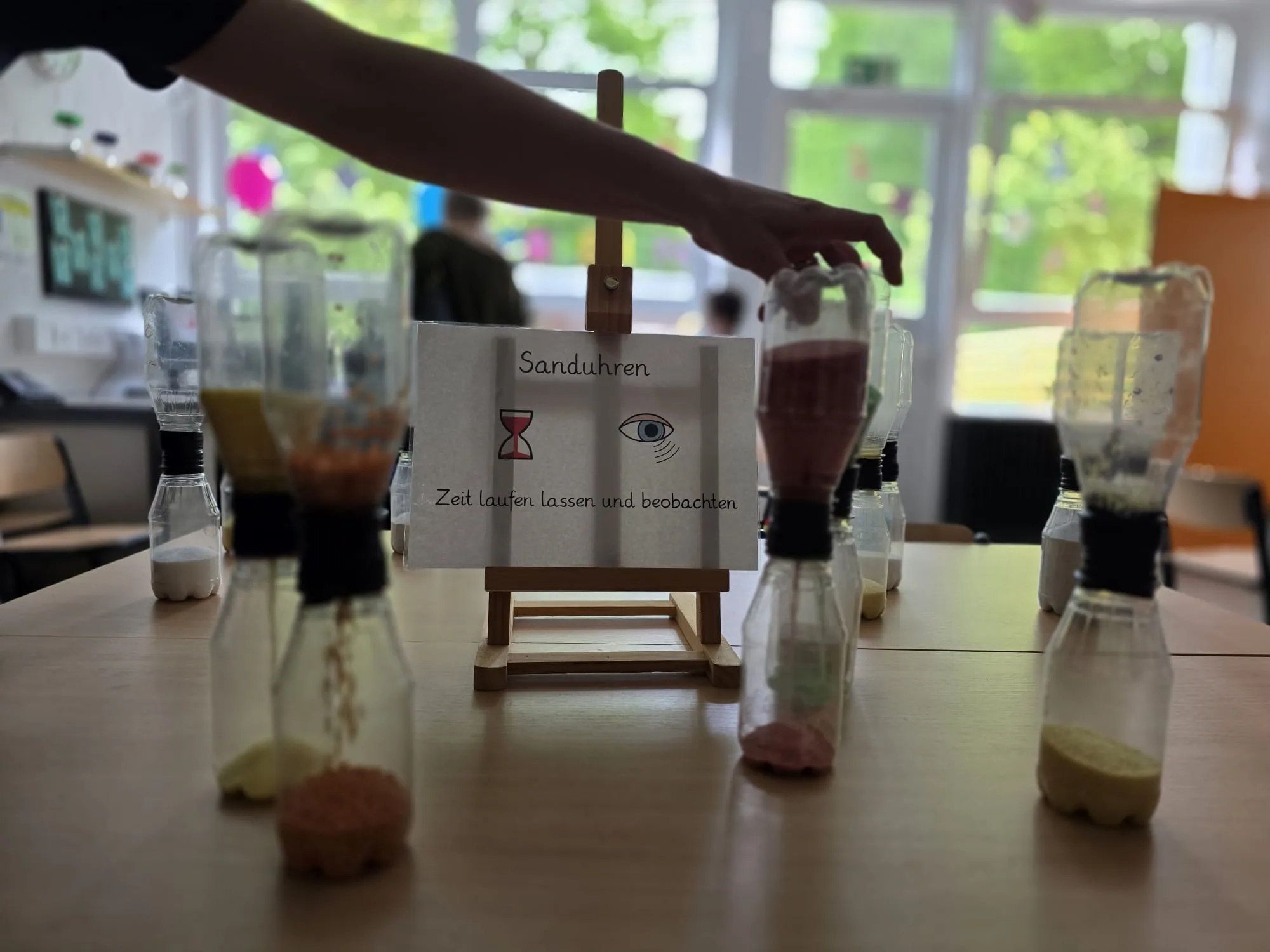 Ein Tisch mit mehreren durchsichtigen Behältern, gefüllt mit unterschiedlich gefärbten Flüssigkeiten und Materialien, steht im Fokus. Davor ein Schild mit der Aufschrift „Sanduhren“ und der Aufforderung, Zeit laufen zu lassen und zu beobachten.