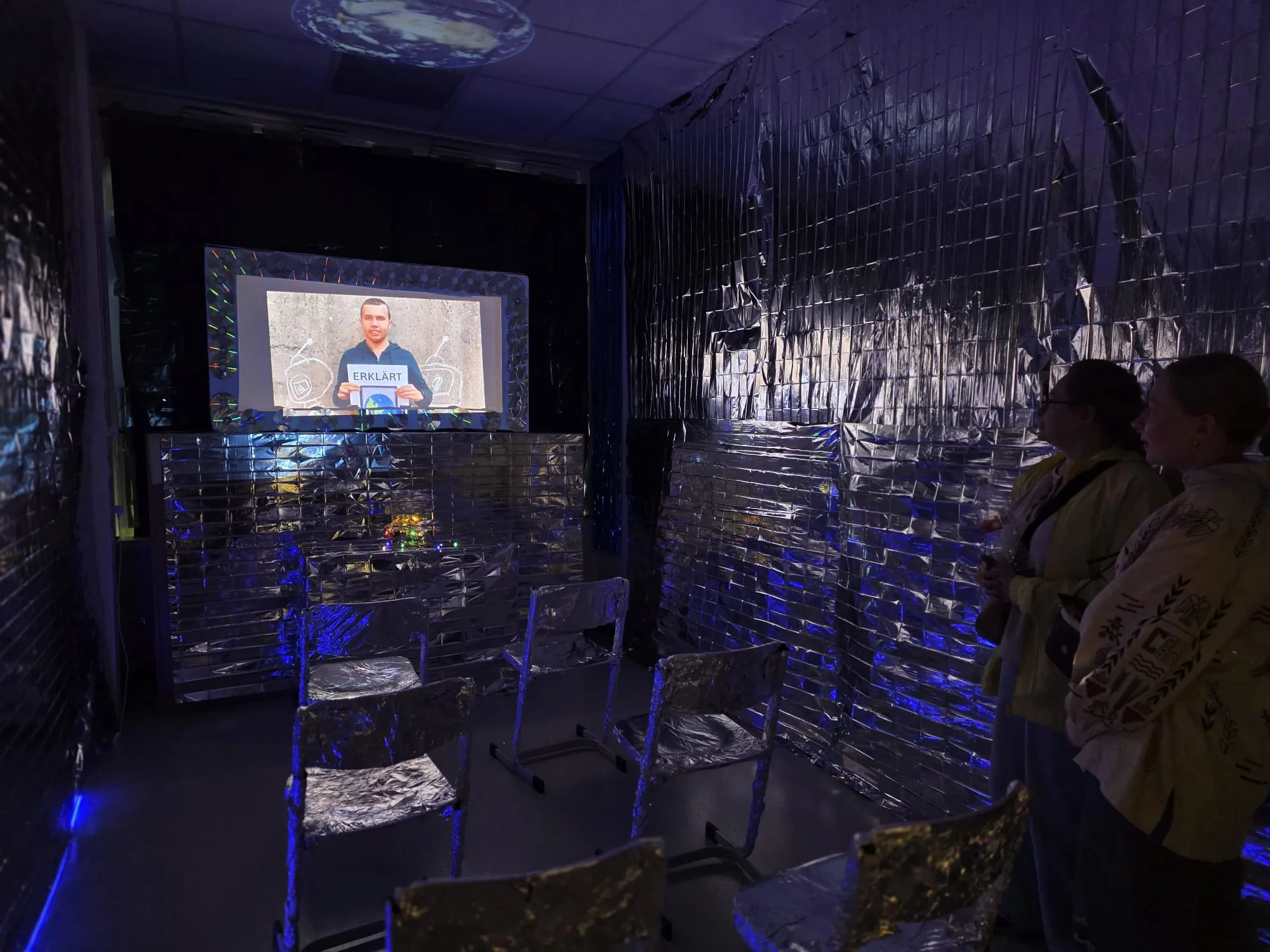 Ein Klassenzimmer ist mit silbernen Folienbahnen verkleidet und wird durch violettes Licht beleuchtet. An der Stirnwand wird auf einer Leinwand ein Mann beim Sprechen gezeigt, während zwei Personen rechts im Bild stehen und zusehen.