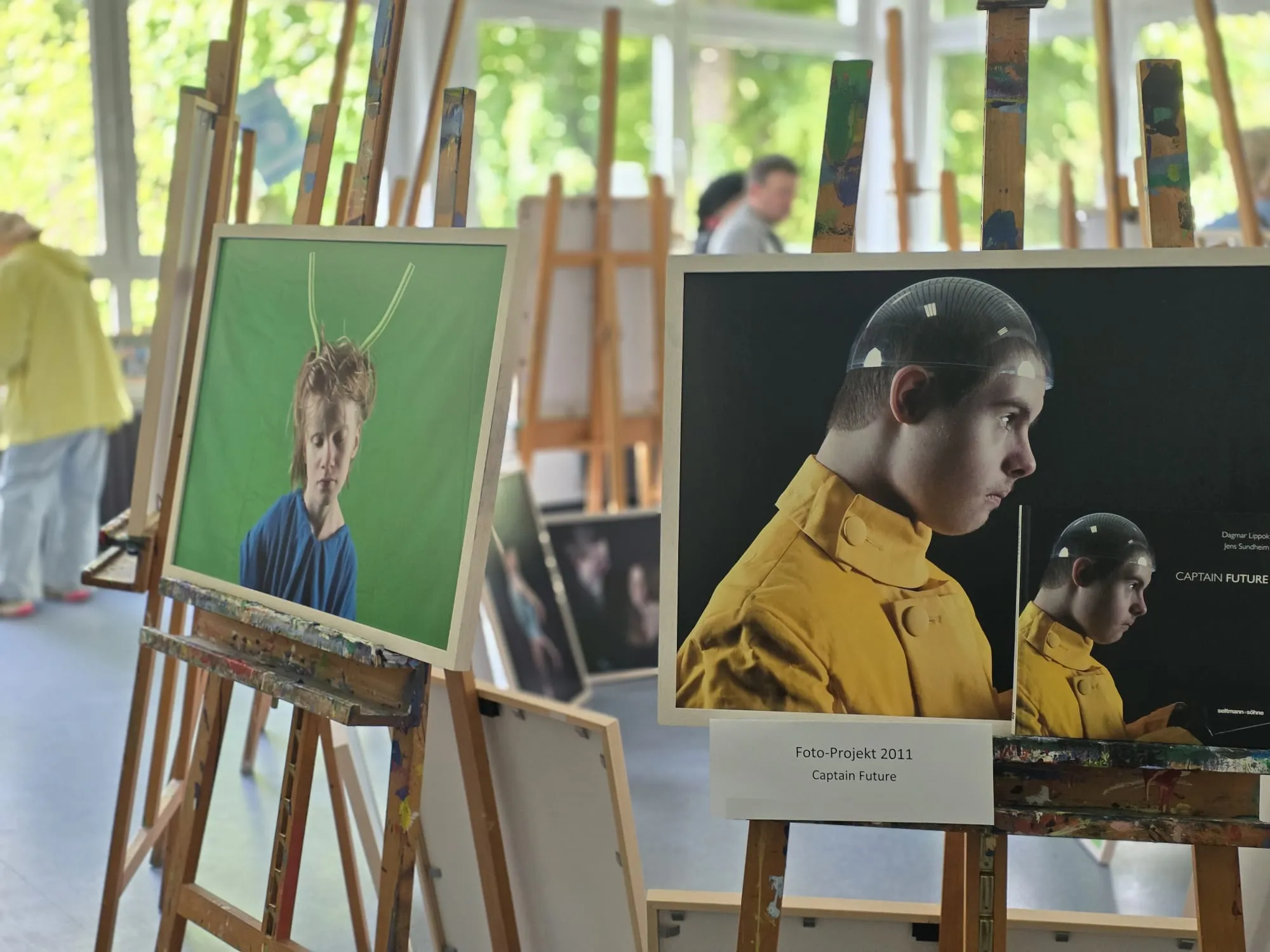 Gemälde und Fotografien stehen auf Staffeleien in einer hellen Ausstellungshalle, im Hintergrund sind weitere Besucher zu sehen.