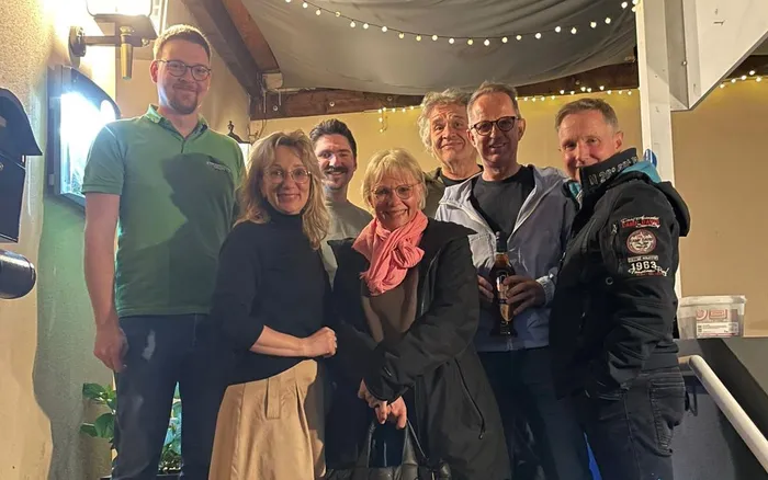 Eine Gruppe von sechs Personen steht lächelnd zusammen. Zwei Männer, Felix Goseberg und Steffen Wieland, sind erkennbar, während ein Mann in der Mitte ein Bier hält.