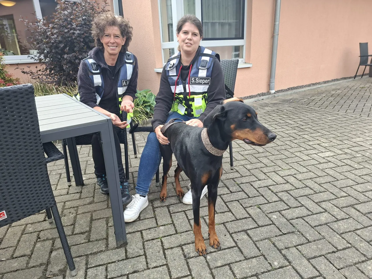 Zwei Frauen in Uniform sitzen an einem Tisch im Freien, neben ihnen steht ein Dobermann. Die linke Frau lächelt in die Kamera, die rechte blickt geradeaus.