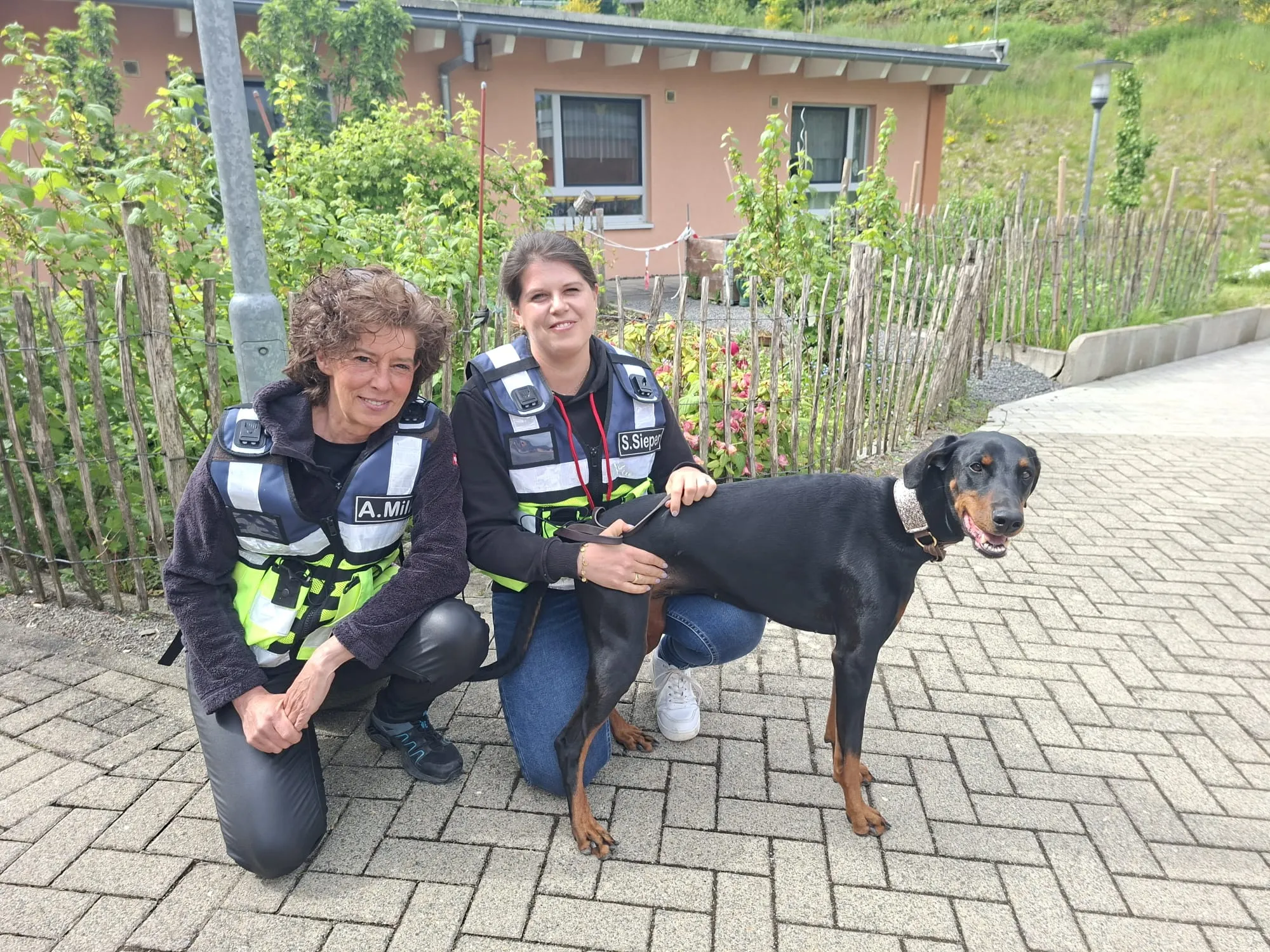 Zwei Frauen in Westen und eine Dobermannhündin posieren auf einem gepflasterten Weg vor Büschen und einem Gebäude. Die Hündin trägt ein Halsband.