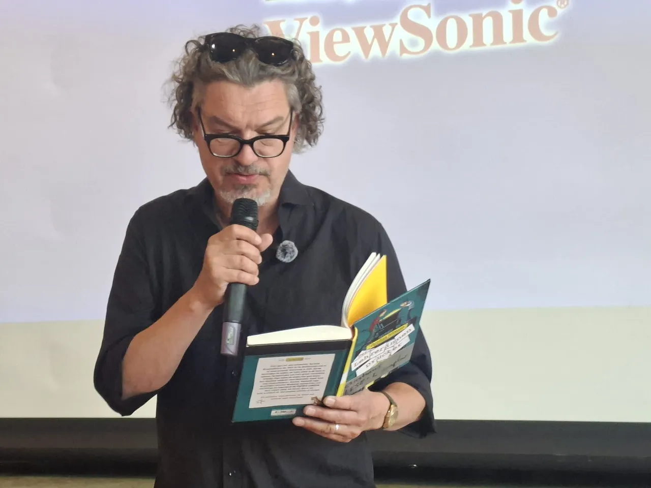 Christian Seltmann liest aus seinem Buch „Der Lehrerinnendrucker“ vor Publikum. Er hält ein offenes Buch in der Hand und spricht in ein Mikrofon.