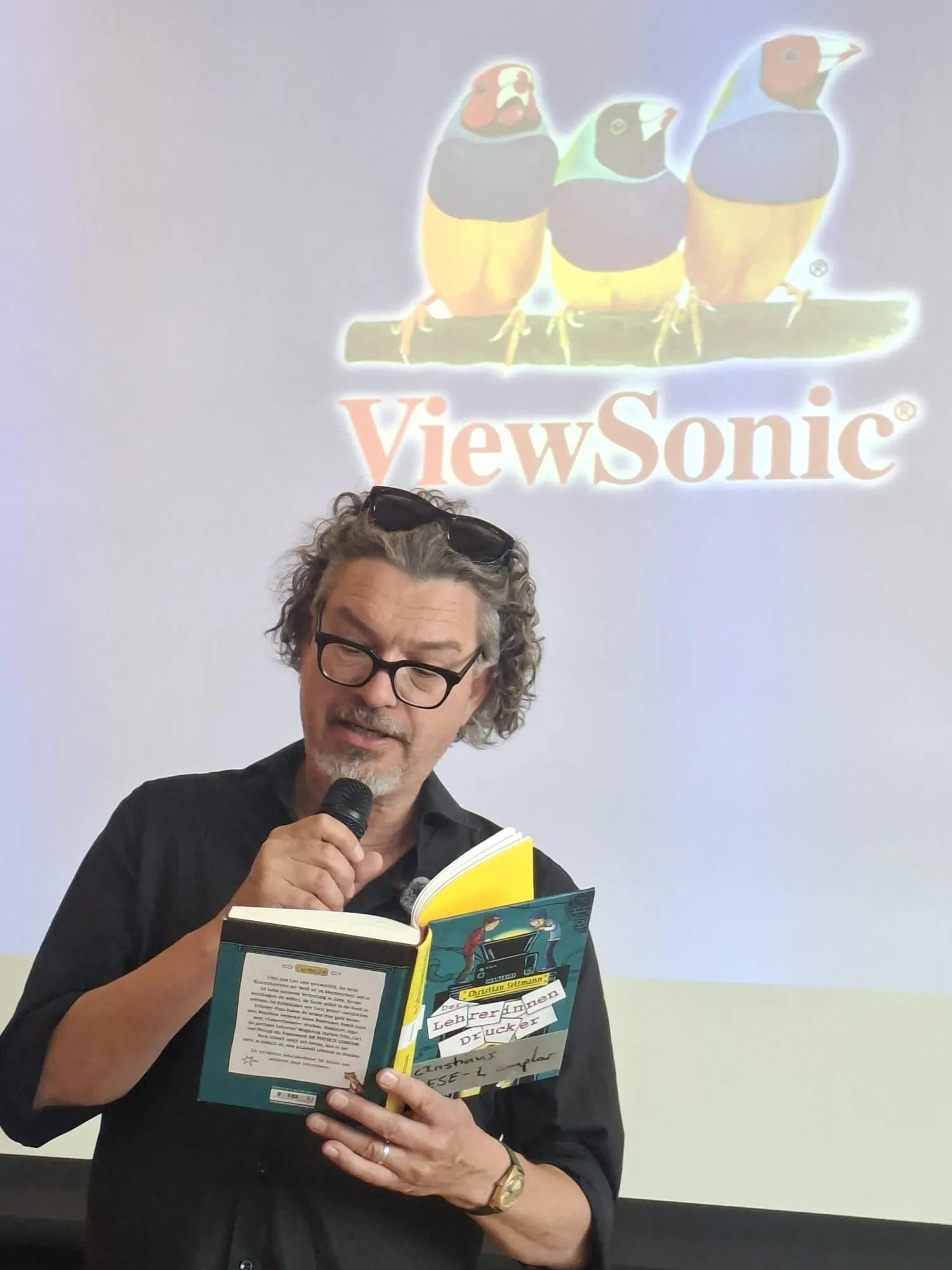Christian Seltmann liest aus seinem Buch „Der Lehrerinnendrucker“ vor Publikum. Im Hintergrund eine Projektion mit dem ViewSonic-Logo und stilisierte Vögel.