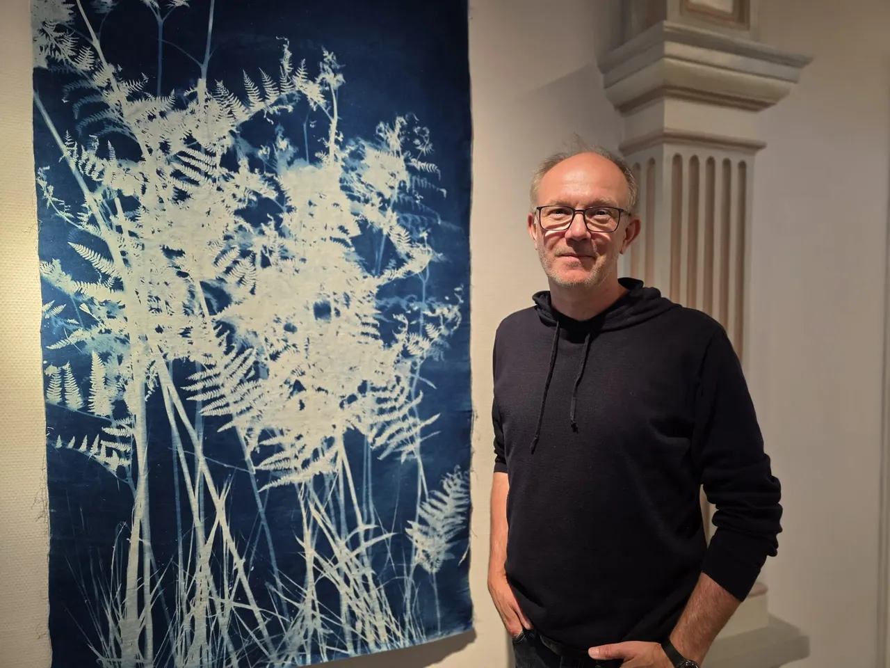 Ein Mann mit Brille und dunklem Hoodie steht neben einem großen, blauen Bild mit Pflanzenmotiven in einer hellen Galerie.