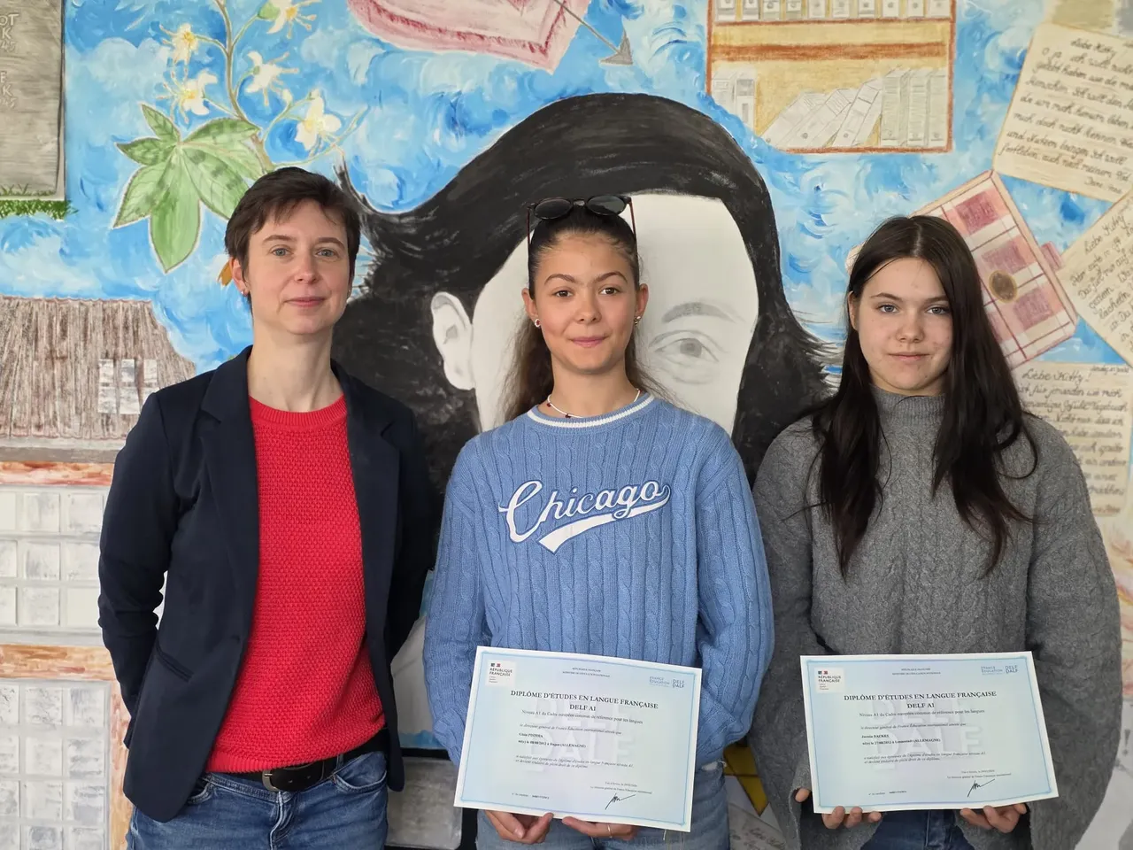 Drei Personen posieren vor einer bunten Wand mit Porträt und Text. Links eine Frau in einem schwarzen Blazer, in der Mitte und rechts zwei junge Mädchen, die jeweils ein Zertifikat in der Hand halten.